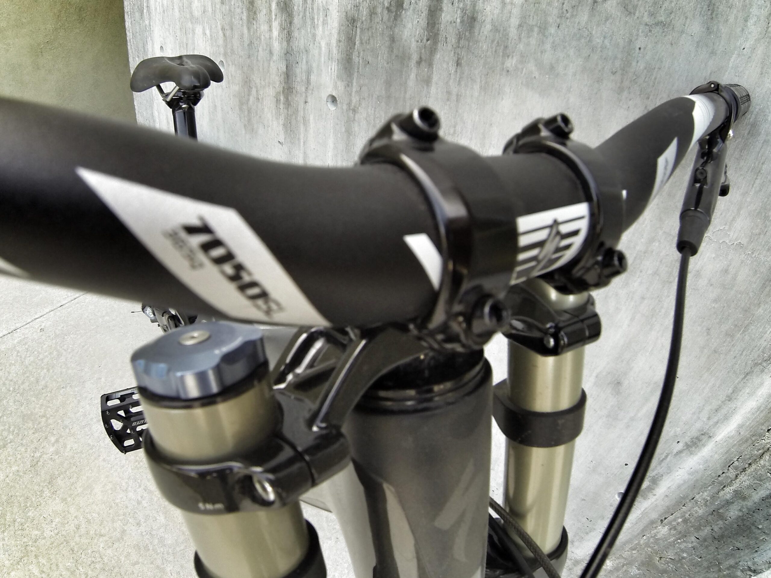 Specialized S-Works Demo 8: Close-up view of a mountain bike's handlebars and front suspension, featuring a dark color scheme, and prominent components including a textured handlebar grip and a suspension adjustment knob. The background shows a plain gray concrete surface.