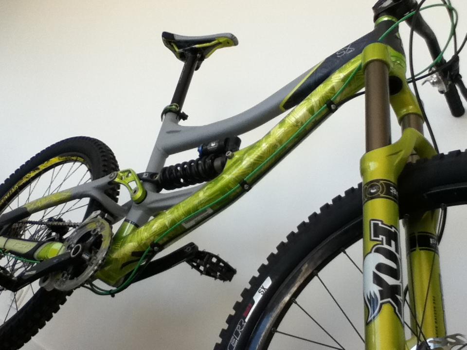 Alt text: "Close-up view of a mountain bike leaning against a wall, featuring a gray frame with green and black accents, a visible suspension system, and knobby tires."