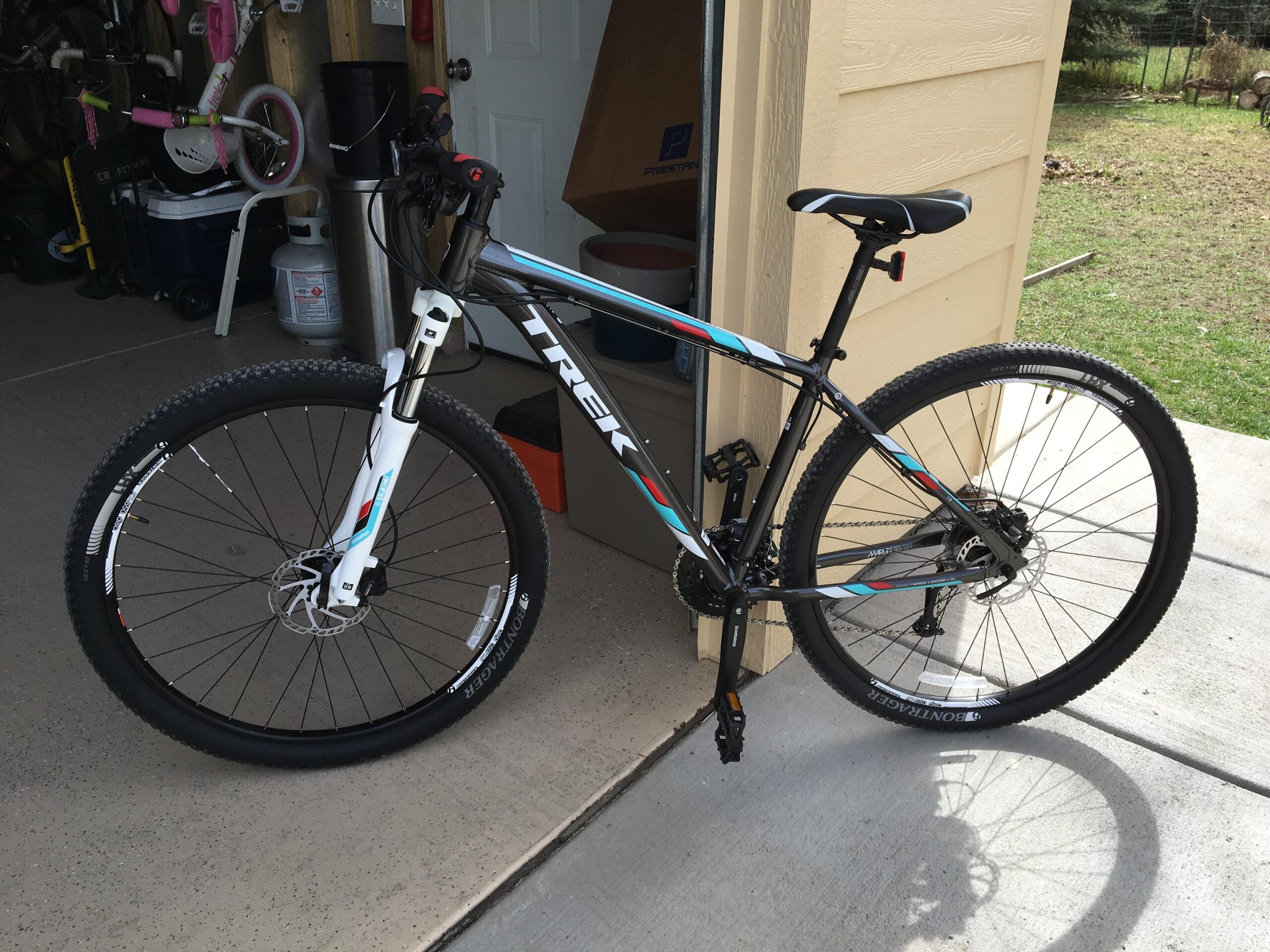 A Trek mountain bike is parked in a garage, showcasing its sleek design and vibrant colors. The bike features a white and blue frame with black accents, disc brakes, and knobby tires suitable for off-road use. In the background, a children's bicycle can be seen, along with various items stored in the garage.