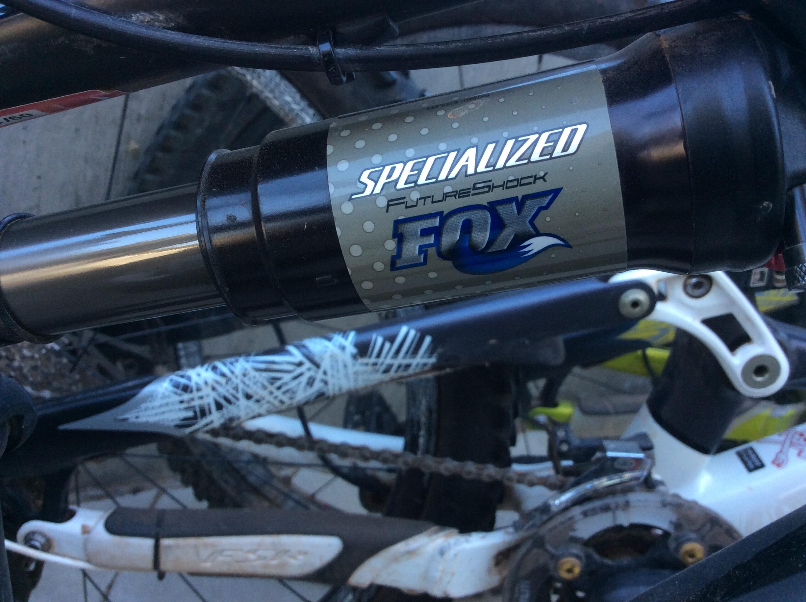 An image showing a close-up of a Specialized FutureShock FOX suspension component attached to a mountain bike frame, with visible bike parts and a textured background.