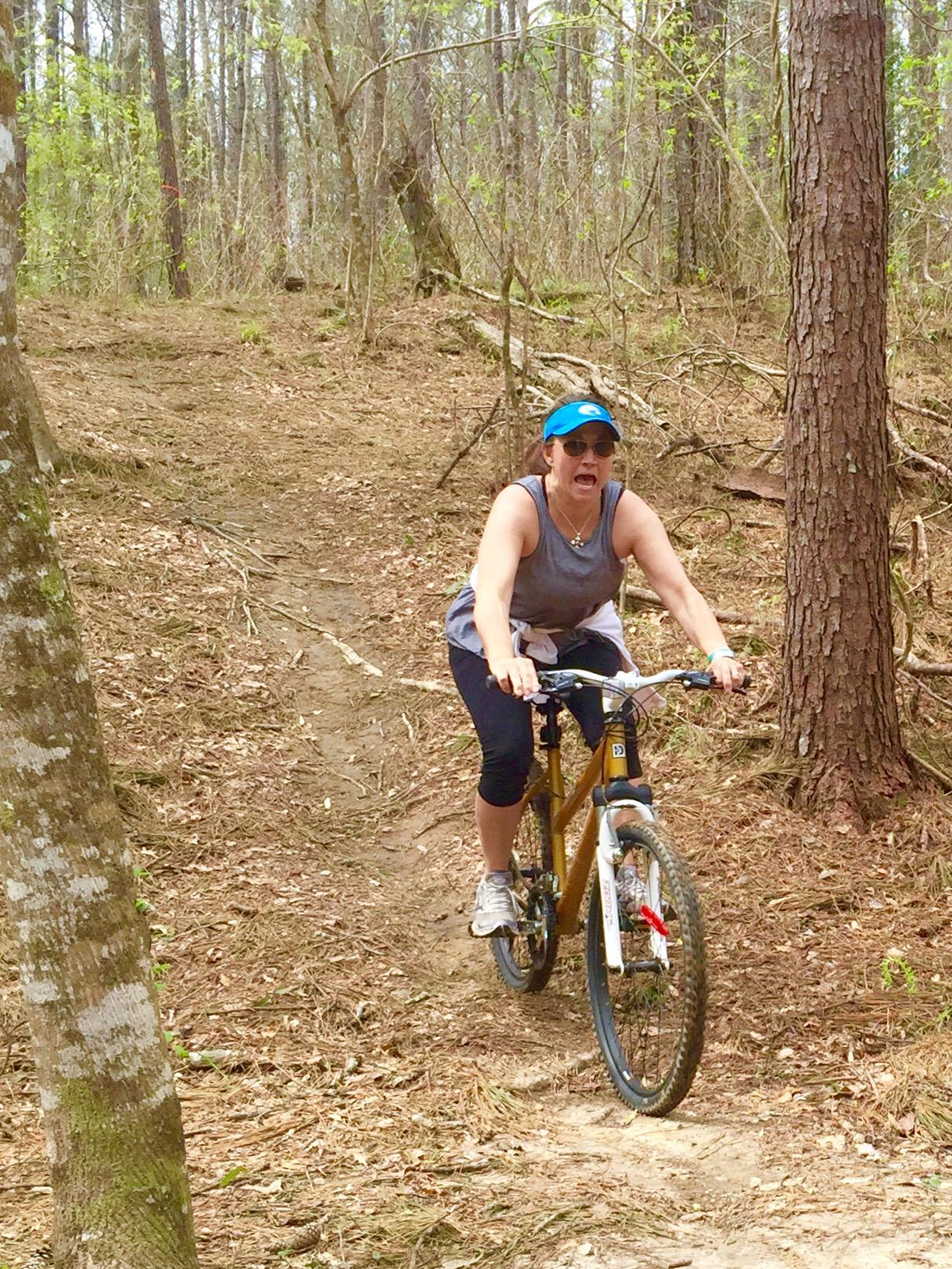 A person riding a mountain bike on a trail in a wooded area, surrounded by trees and foliage. They appear to be engaging with the terrain, wearing a tank top, sunglasses, and a cap. The trail is slightly uphill, and the ground is covered with leaves and dirt. Mt. Zion Bike Trails mountain bike trail.