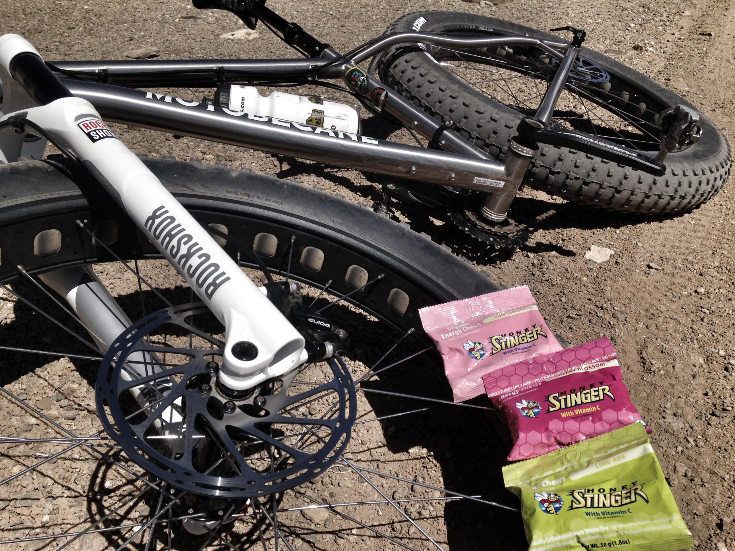 Motobecane Motobecane Bullet Titanium Fat bike: A close-up view of a mountain bike resting on dirt, featuring a prominent white front fork and a disc brake system. Next to the bike, three packets of Honey Stinger energy chews in different flavors are displayed, showcasing vibrant packaging.