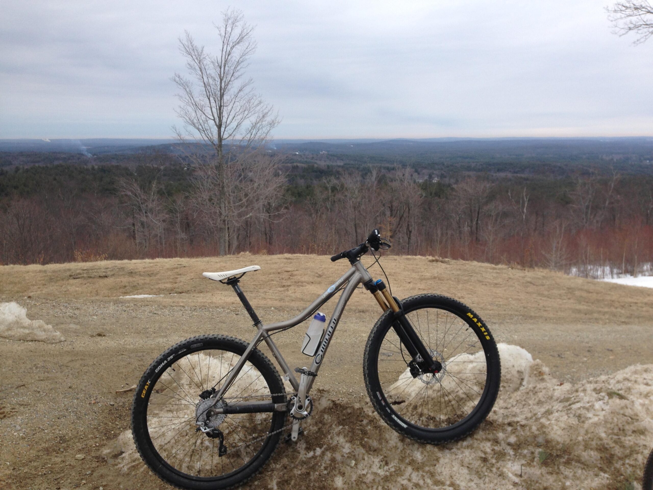 Canfield Brothers Yelli Screamy: A mountain bike resting on a snowbank, overlooking a scenic view of rolling hills and a cloudy sky in the background. The landscape features a mix of bare trees and patches of grass, indicating early spring or late winter conditions.