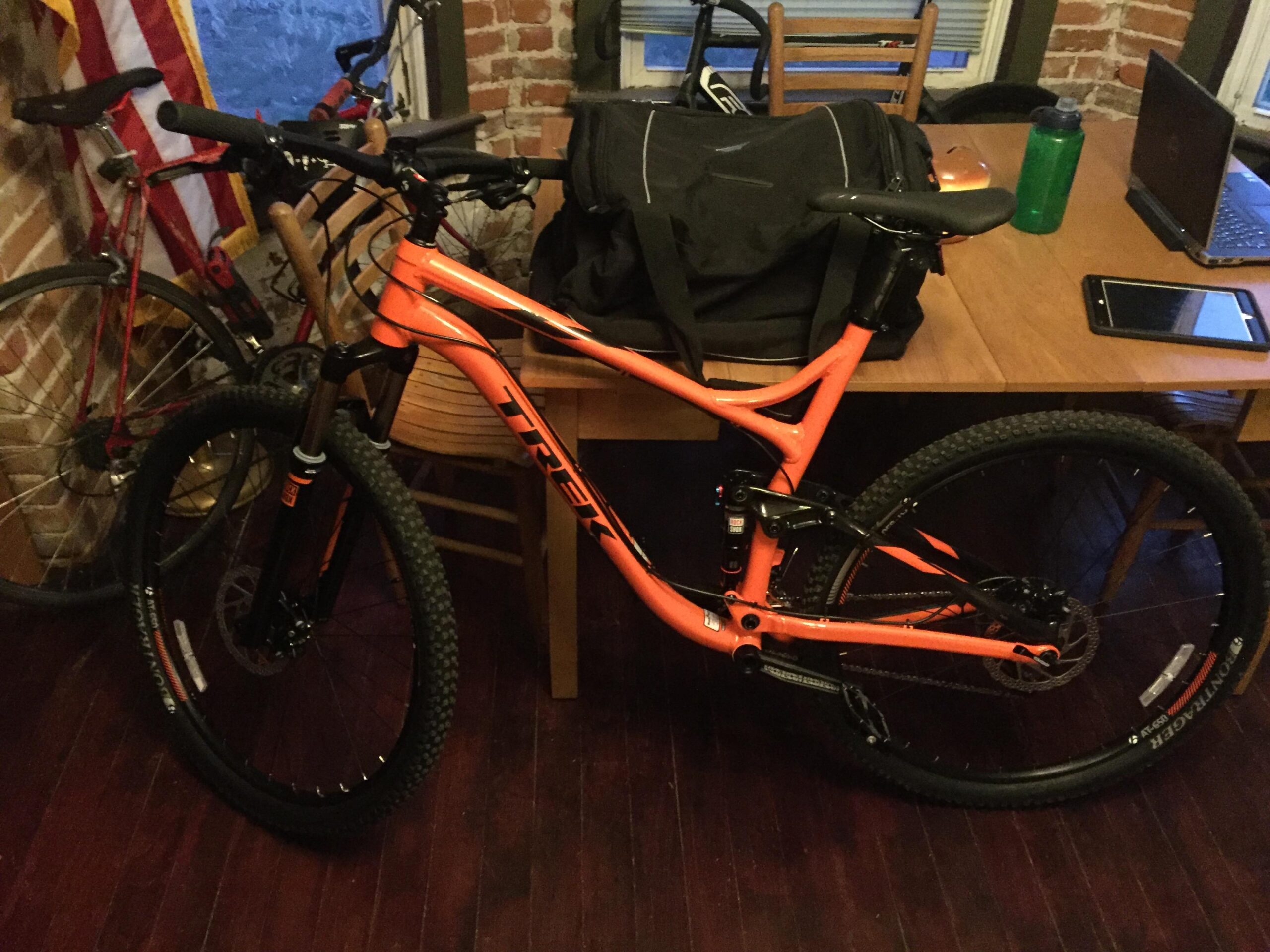 Trek Fuel EX 5 29: A vibrant orange Trek mountain bike is leaning against a wooden table in a room. Behind it, a red bicycle is partially visible, and various items like a black bag, a green water bottle, and a laptop are on the table. The space features exposed brick walls and wooden flooring.