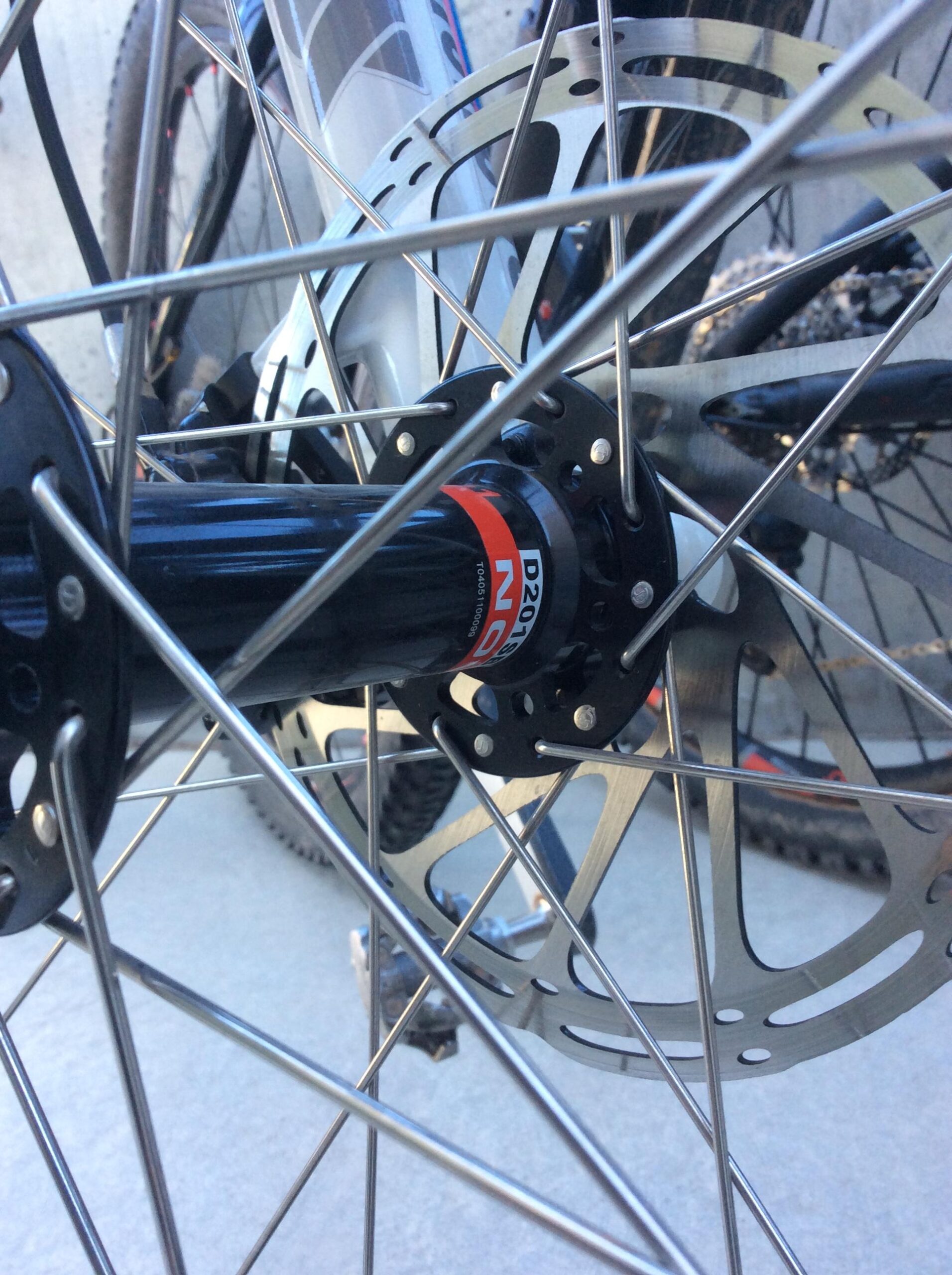 Close-up view of a bicycle wheel hub, showcasing the black hub with a red label and silver disc brake rotor, surrounded by metal spokes and part of the wheel's frame.