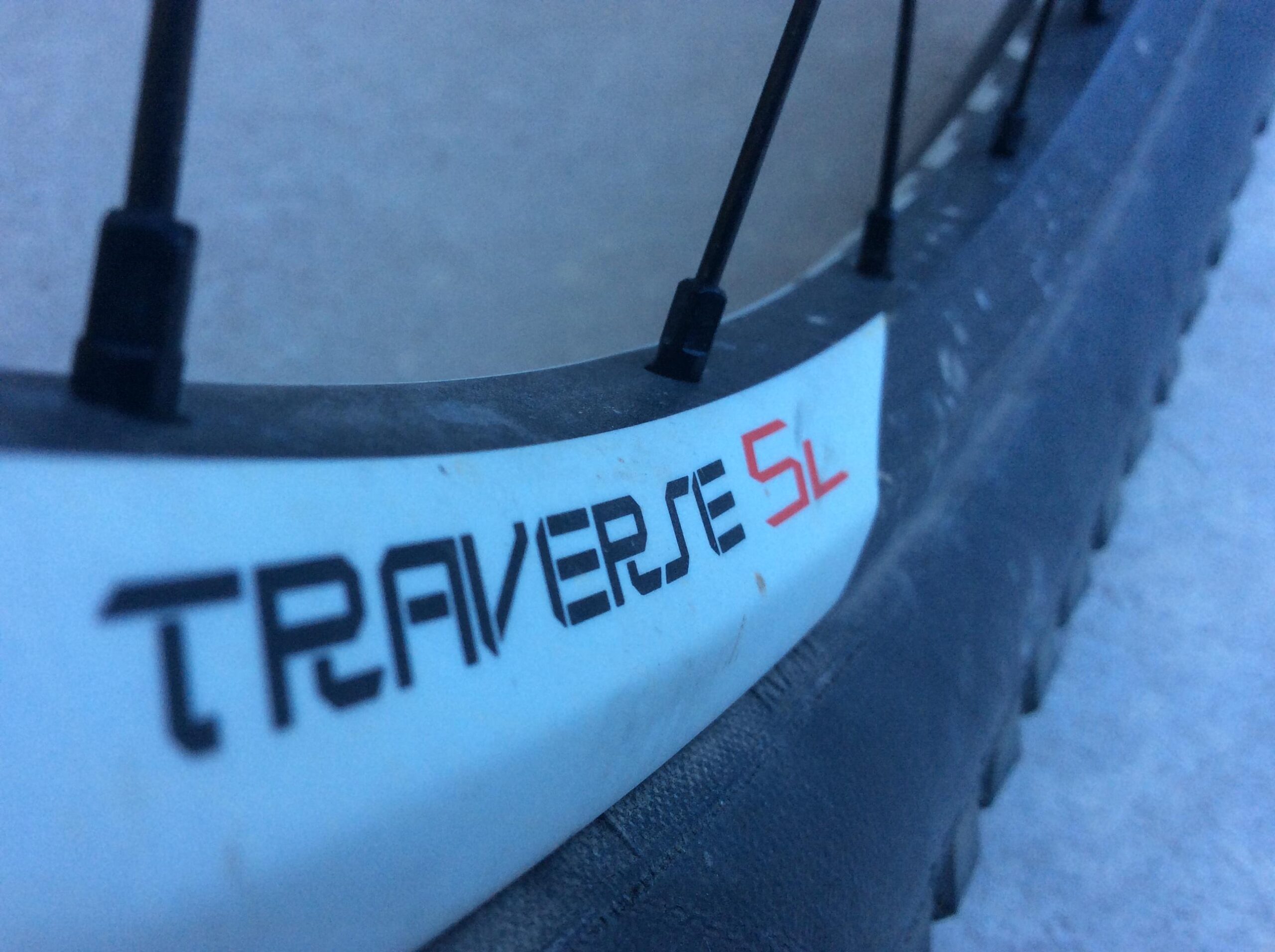 Close-up image of a bicycle wheel featuring the label "TRAVERSE 54" on the rim, with visible spokes and a partially worn tire. The background is blurred and appears to be a light surface.