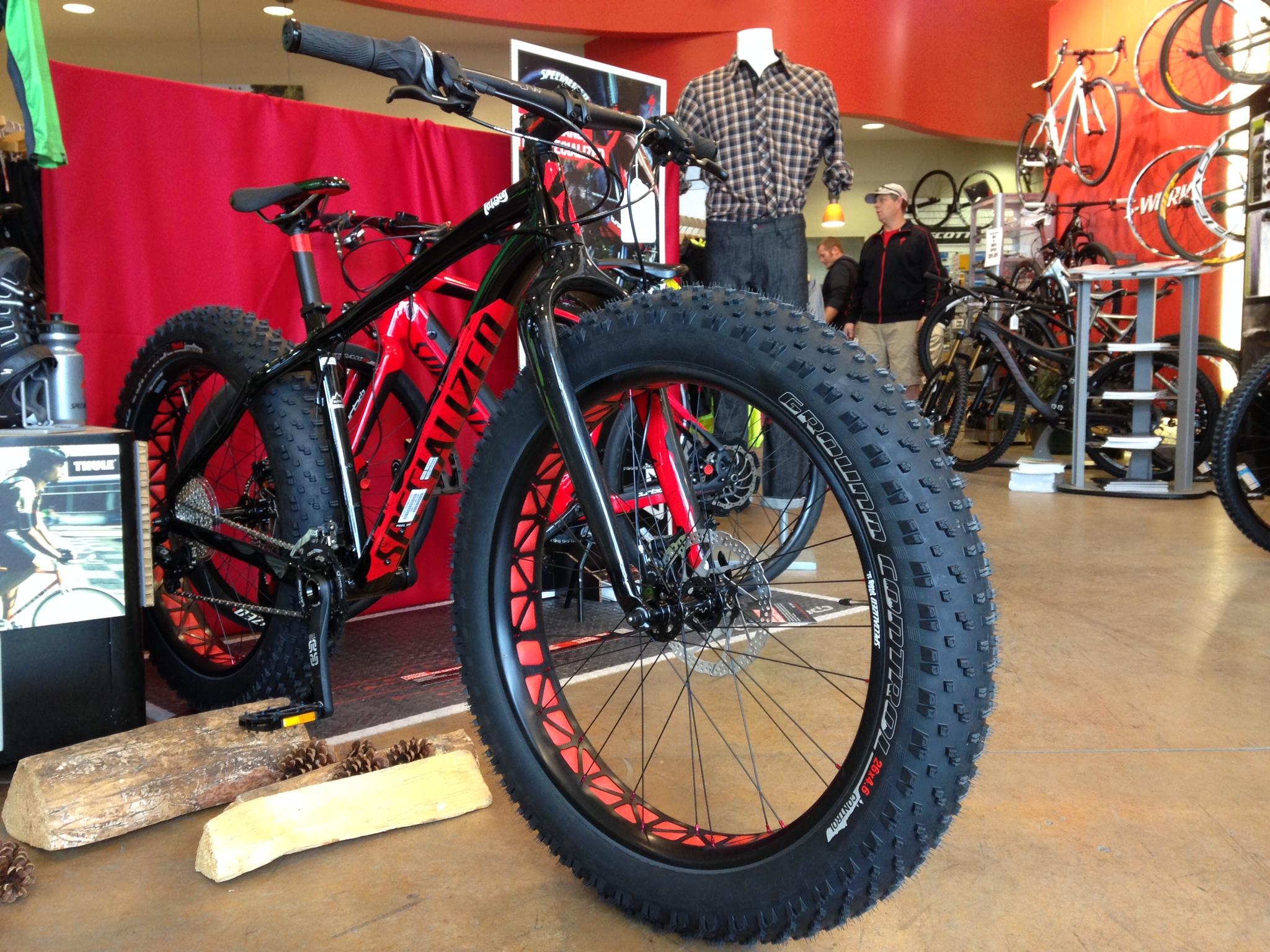 A fat tire bike with a black frame and red accents is prominently displayed in a bike shop. The bike features wide, knobby tires and is positioned near wooden logs and pine cones. In the background, other bicycles and apparel are visible, along with customers browsing the store.
