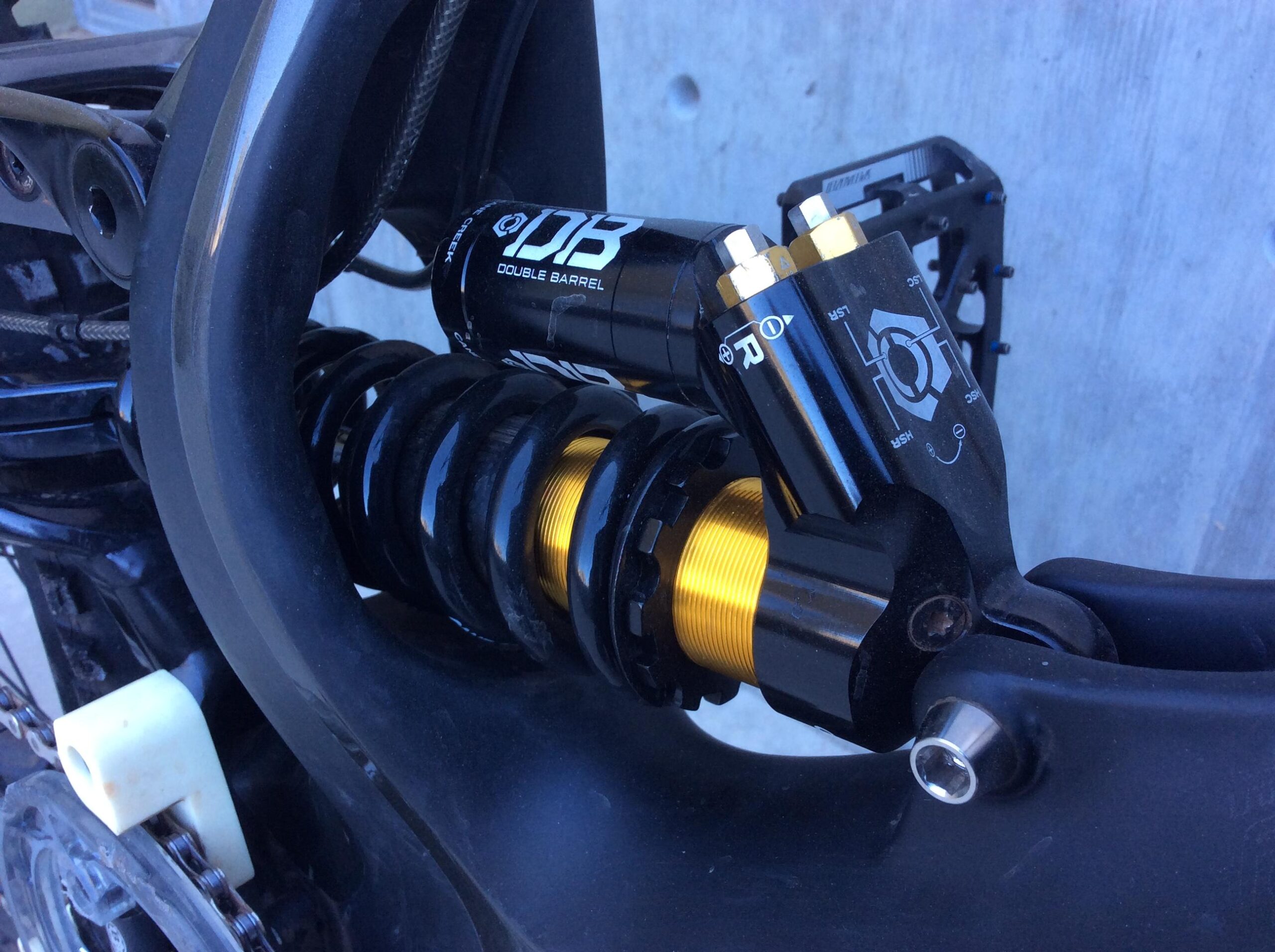 Specialized S-Works Demo 8 Carbon: Close-up view of a mountain bike suspension system, featuring a black and gold shock absorber labeled "Double Barrel." The image shows the compressed spring and the adjustment knobs on the shock, emphasizing the intricate mechanics of the suspension setup. A portion of the bike frame is visible in the background.