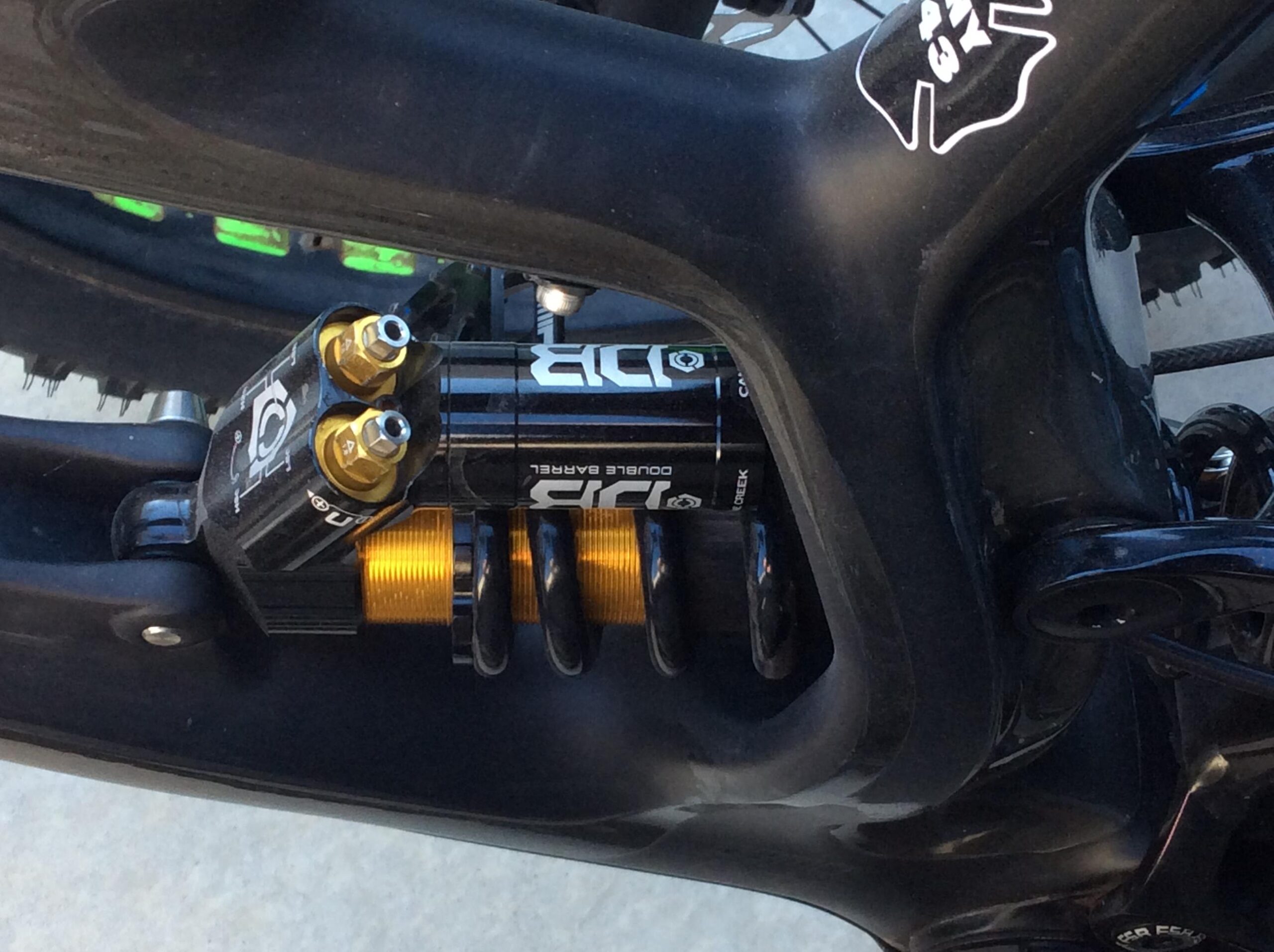 Close-up view of a bike suspension system, featuring a black and gold shock absorber with dual barrels. The shock is mounted within the bike frame, showcasing its intricate design and components. The background shows part of a treaded tire, indicating a mountain biking setup.