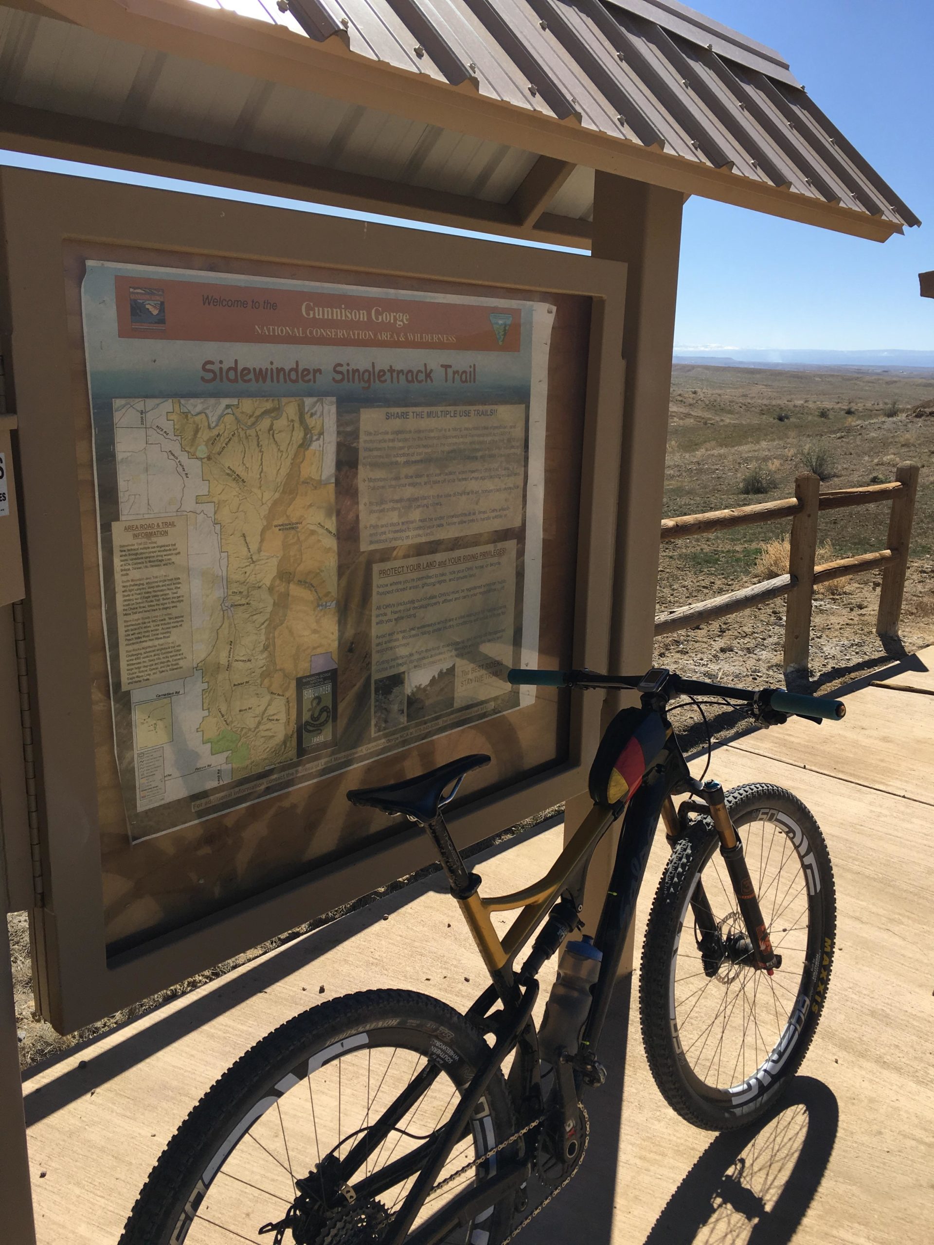 A mountain bike is parked next to a trailhead sign for the Sidewinder Singletrack Trail, located in Gunnison Gorge National Conservation Area. The sign features a map and information about the trail, with a clear blue sky and desert landscape in the background. Sidewinder mountain bike trail.