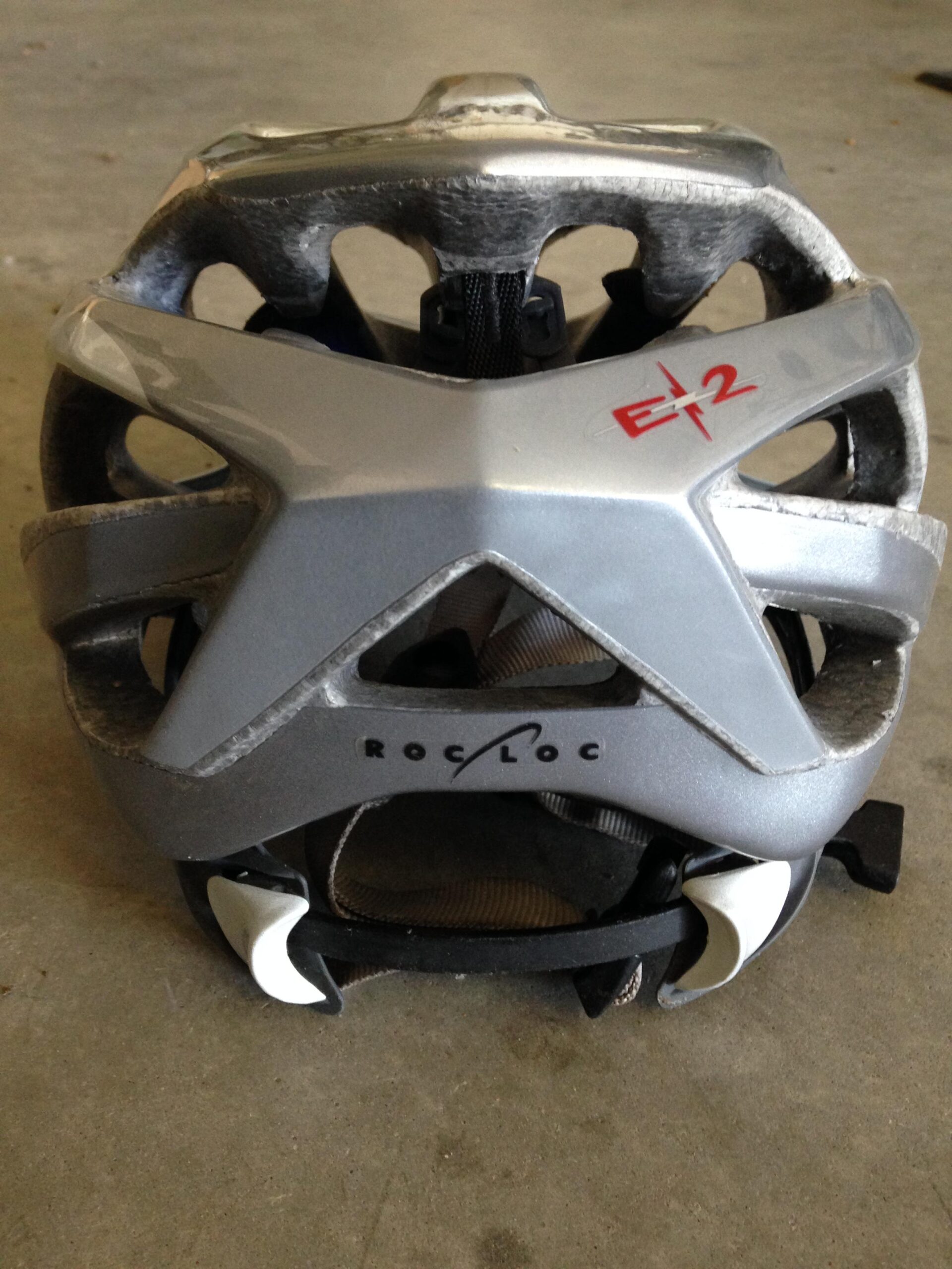 A top view of a silver bicycle helmet with ventilation holes, showing signs of wear and scratches. The helmet features the Roc Loc adjustment system on the back and has the model name "E2" marked in red. The background is a plain, light gray surface.