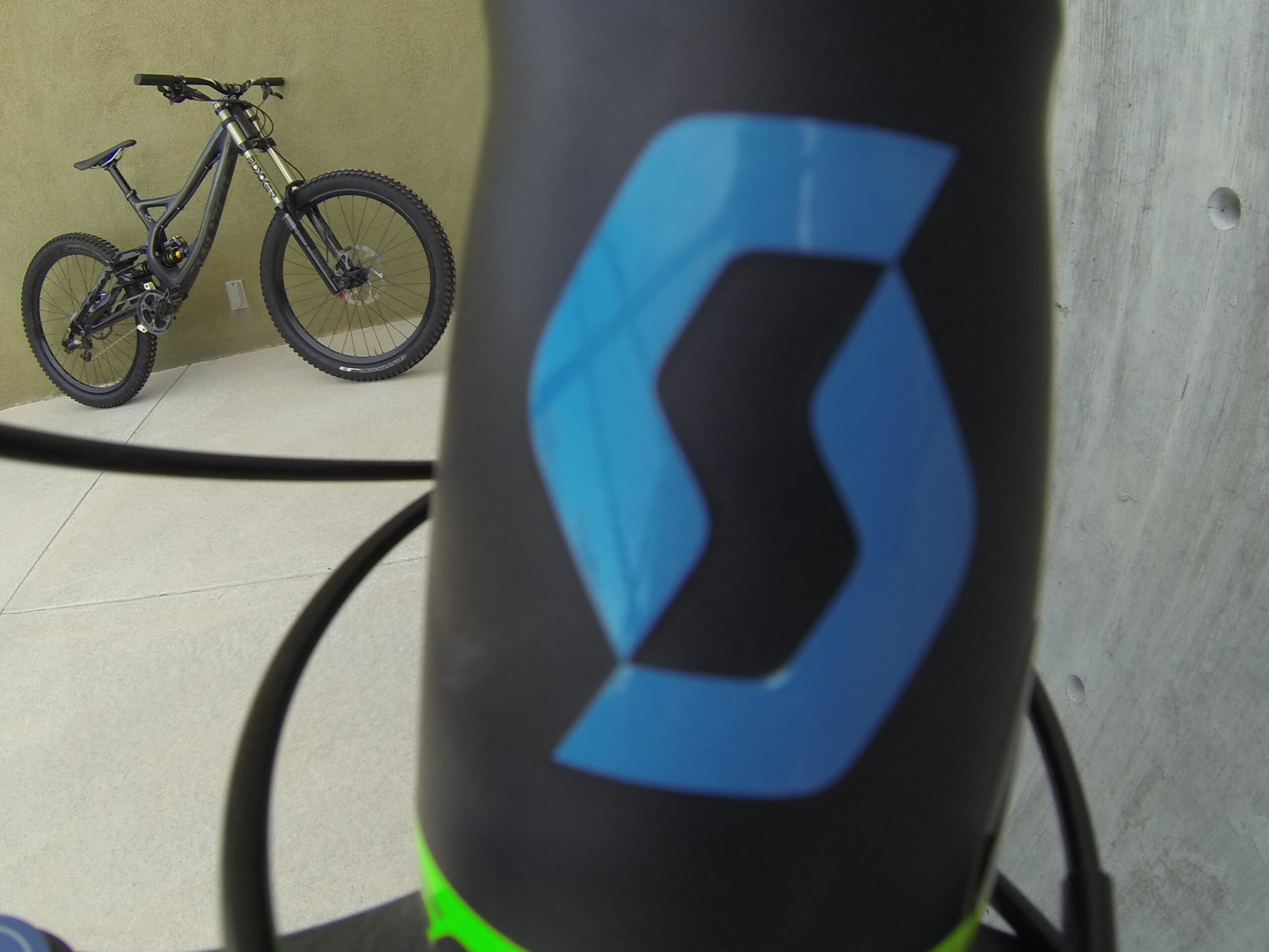 Scott Big Ed: A close-up view of a mountain bike frame featuring a prominent logo, with the bike partially visible in the background against a textured wall. The focus is on the logo, with bike components and cables also visible.