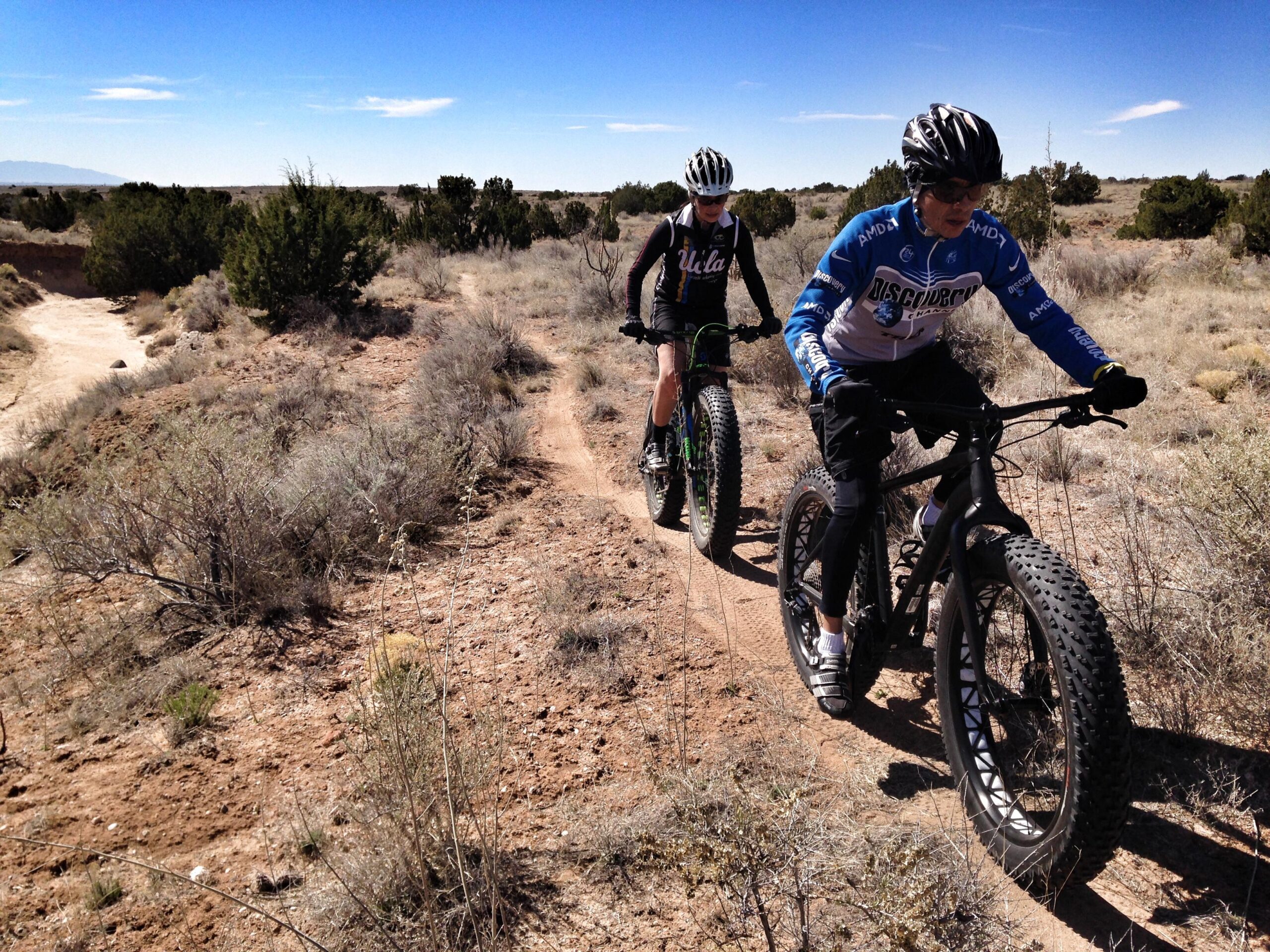 Two cyclists riding fat bikes along a dirt trail in a natural landscape. One cyclist is wearing a blue and white jersey, while the other is in a black jersey. The terrain features sparse vegetation, with shrubs and dry earth under a clear blue sky. Parkway Fatbike trail mountain bike trail.