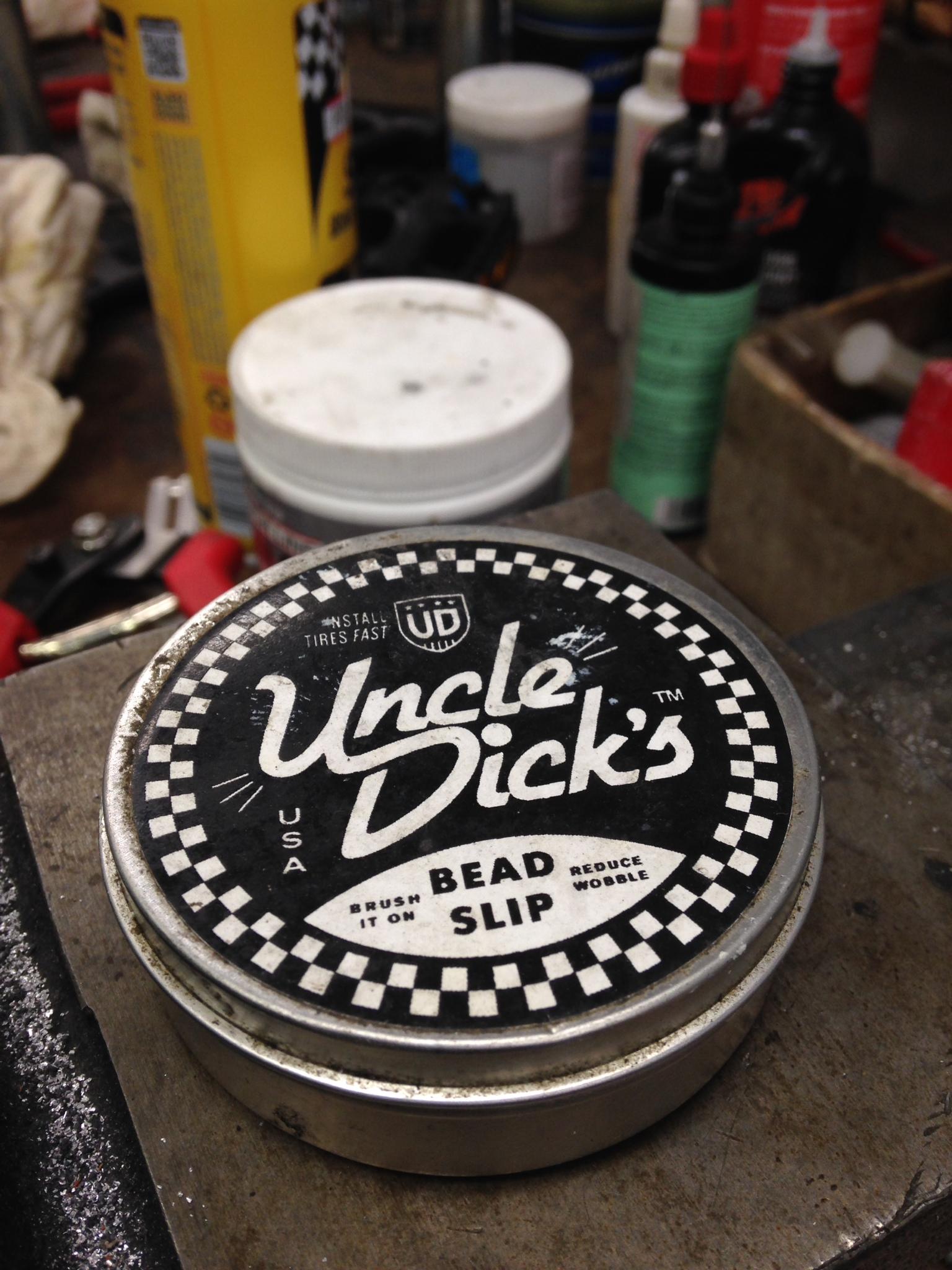 A close-up of a round tin can labeled "Uncle Dick's" with a black and white checkered design, sitting on a workbench cluttered with various tools and supplies. The label indicates it contains a product for tire installation, specifically for reducing wobble and facilitating bead slip.