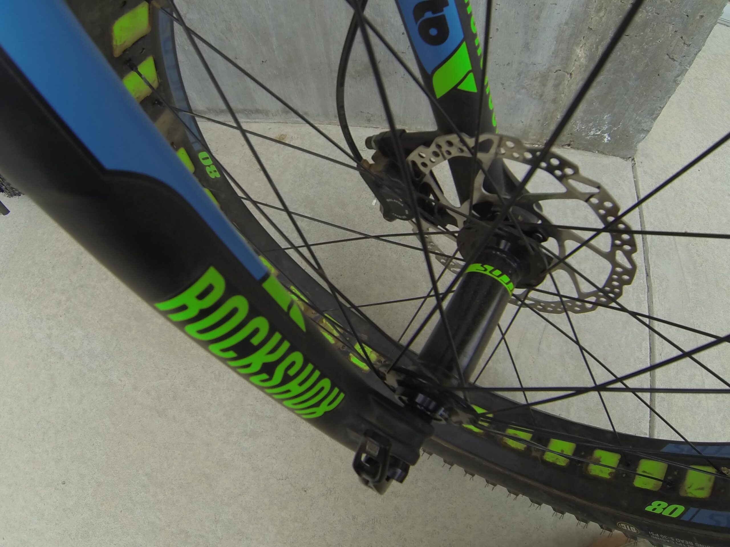 Scott Big Ed: Close-up view of a bicycle front wheel, featuring a black and neon green tire and a visible disc brake system. The fork includes branding for "RockShox," highlighting its suspension features. The background shows a concrete surface.