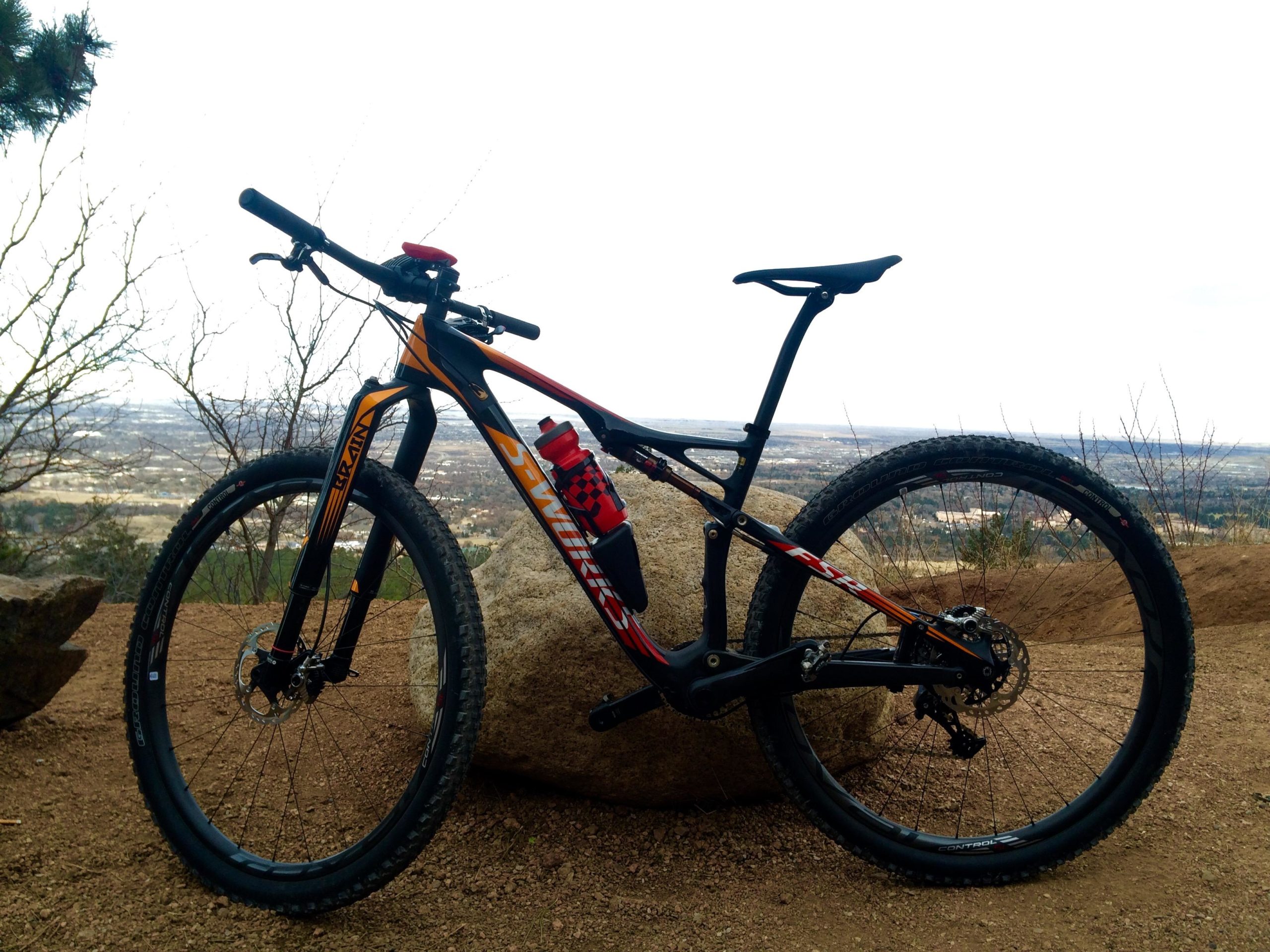 A mountain bike is resting on a rock, with a scenic view of the landscape in the background. The bike features a lightweight frame, thick tires, and a water bottle mounted on its frame. The surroundings include sparse trees and a dirt path, typical of a mountainous terrain. Stratton Open Space / The Chutes mountain bike trail.