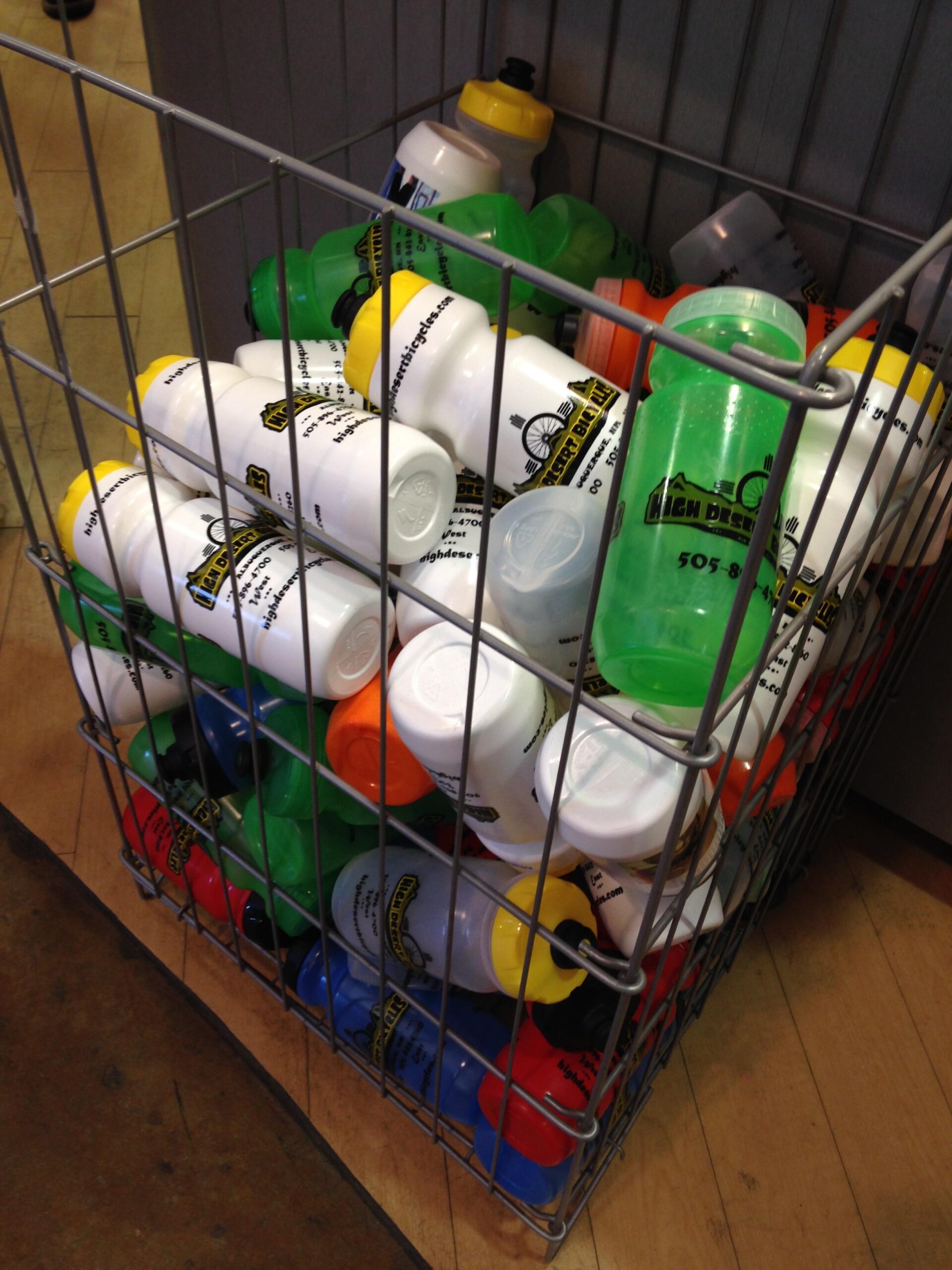 A wire basket filled with various colorful water bottles, including white, green, yellow, red, and blue. The bottles display text and logos, indicating they are branded merchandise from a business. The background shows a wooden floor.