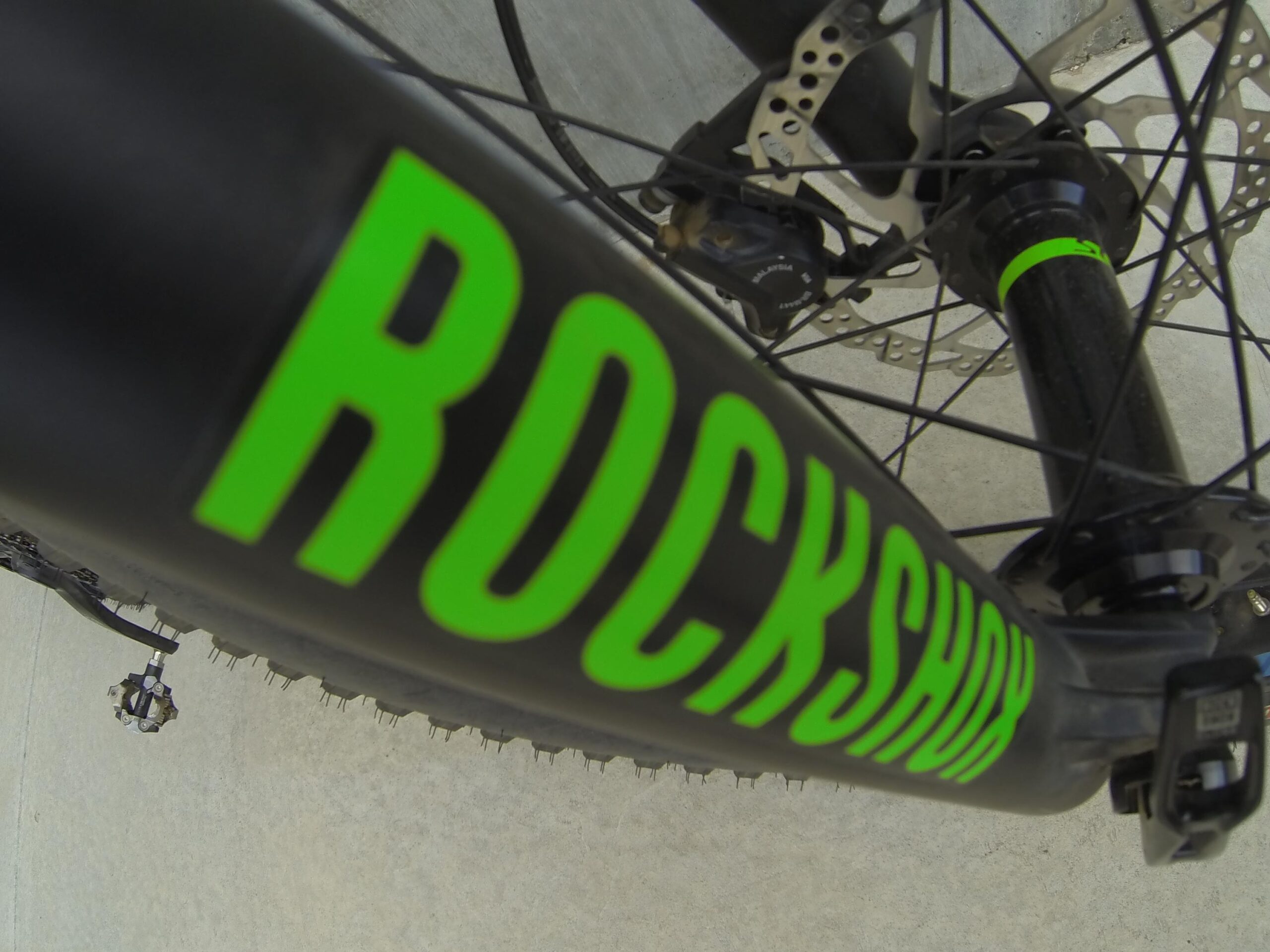 Scott Big Ed: Close-up view of a bicycle fork with the logo "ROCKSHOX" prominently displayed in bright green. The image shows a portion of the bike's wheel, brake components, and pedals, set against a light gray background.