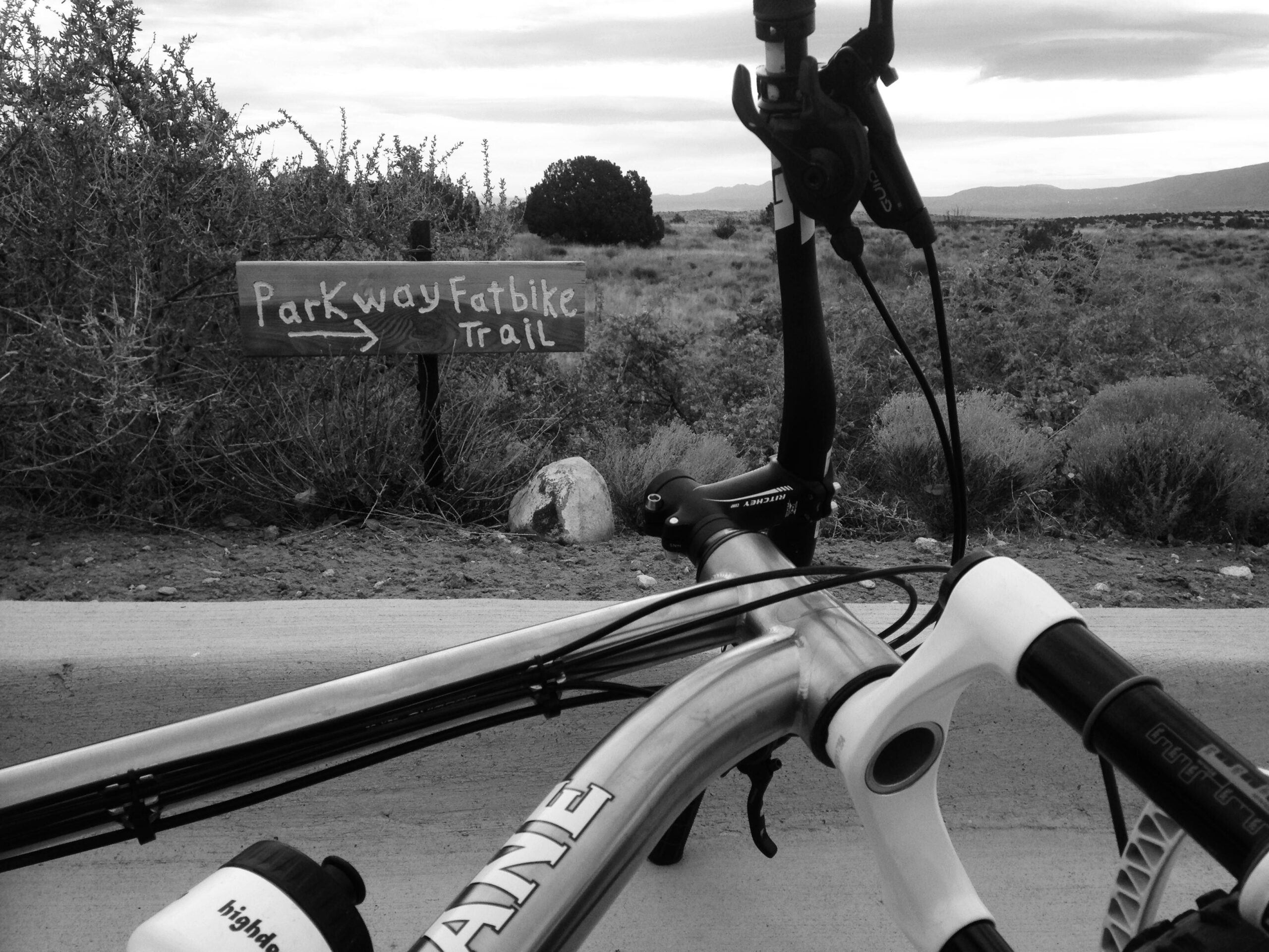 A close-up image of a bicycle's handlebars resting on a path, with a wooden sign in the background indicating the direction to the Parkway Fatbike Trail. The surroundings feature sparse vegetation and rocky terrain under a cloudy sky. The image is in black and white. Parkway Fatbike trail mountain bike trail.