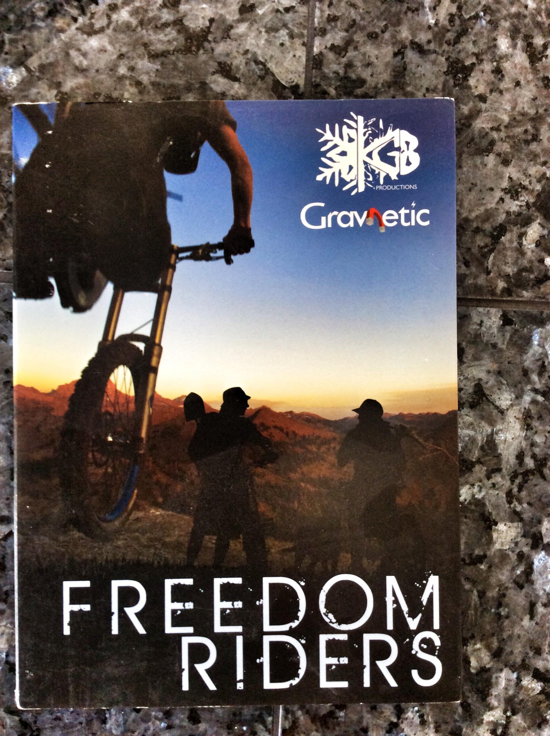 Cover of a video or film titled "Freedom Riders," featuring a silhouette of a mountain biker performing a jump against a sunset backdrop. The design also includes the logos for KGB Productions and Gravnetic. The surface is a textured gray stone.