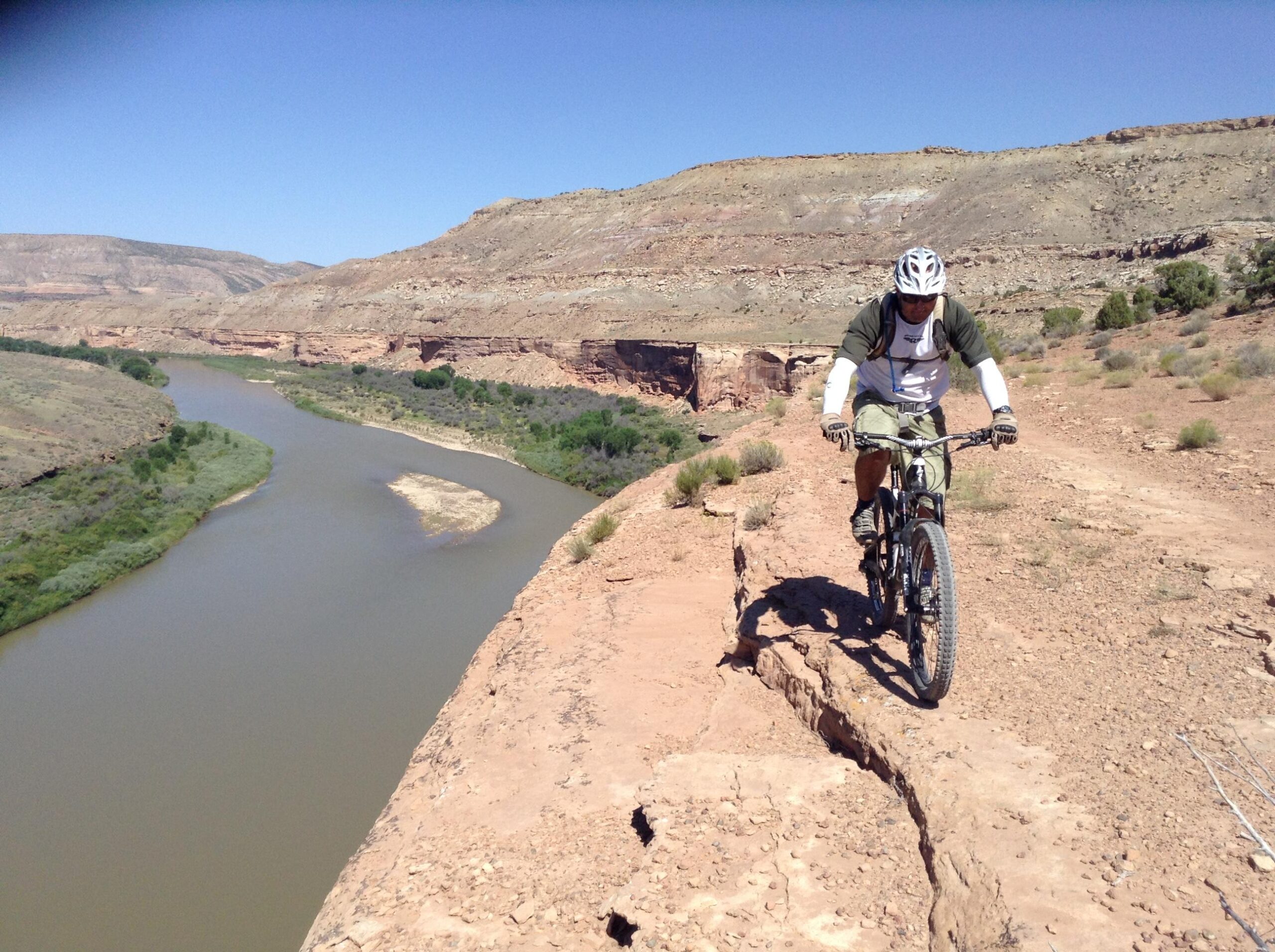 Specialized S-Works Enduro SL Carbon: A mountain biker navigating a narrow rocky path along a cliffside, with a winding river and verdant landscape visible below, set against a clear blue sky and rugged hills in the background.
