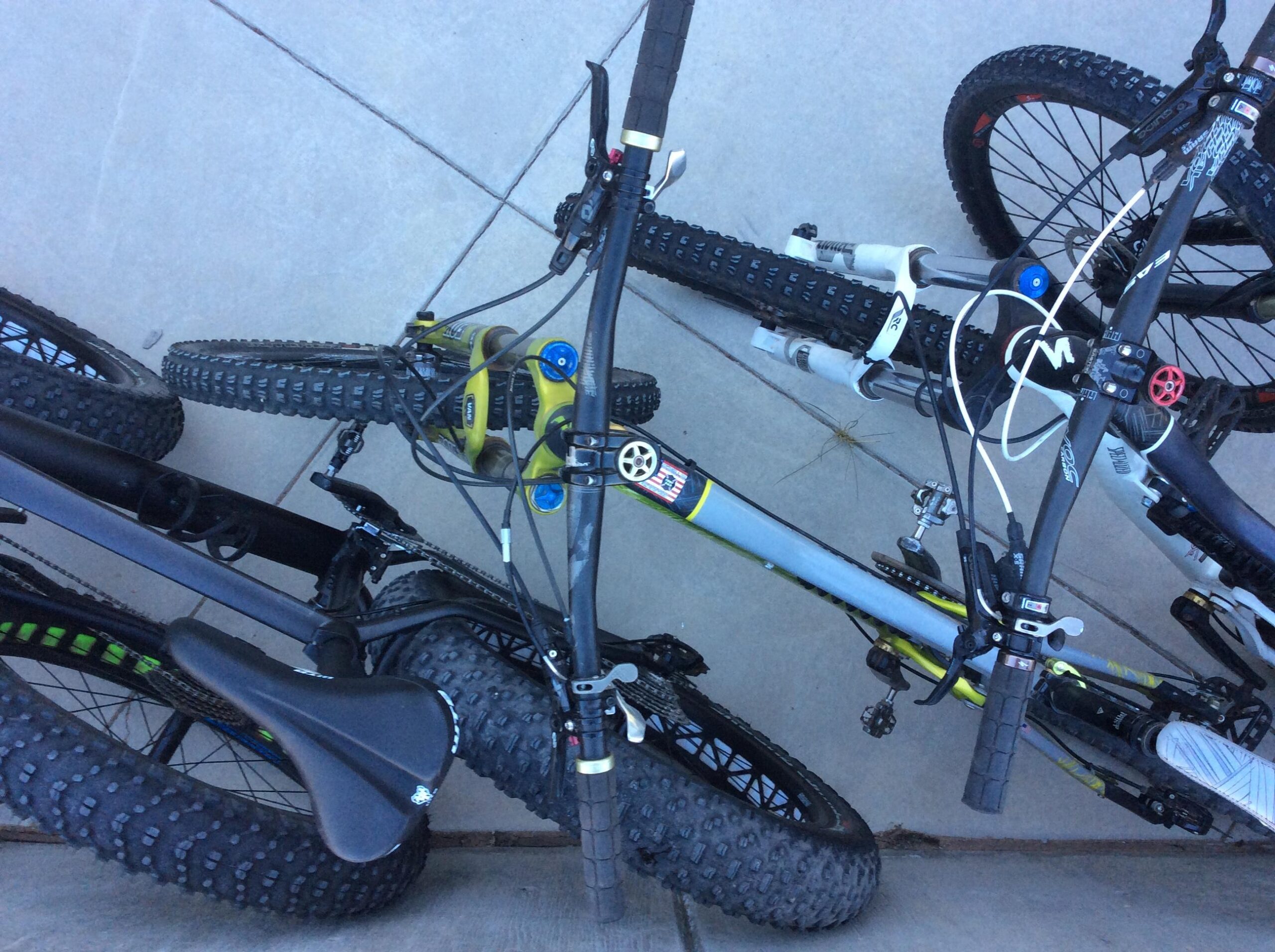 A close-up view of two mountain bikes lying on a concrete surface, showcasing their tires, handlebars, and a mix of components such as grips and saddles. The bikes are intertwined, with visible details like chain and brake cables.