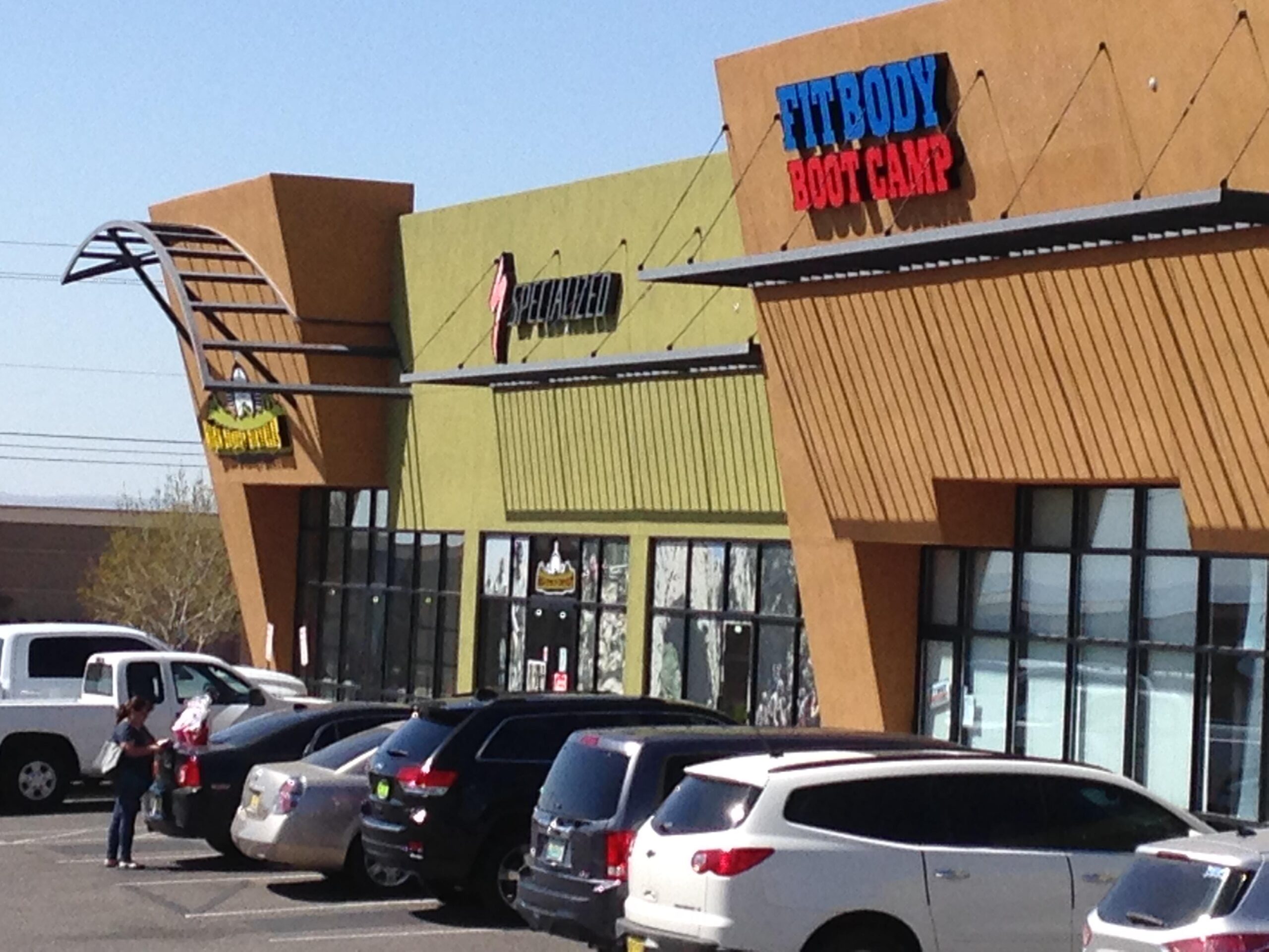 A colorful shopping plaza featuring two storefronts: one with a green exterior labeled "Specialized" and another with a brown exterior labeled "Fit Body Boot Camp." Several parked cars are visible in the foreground, along with a woman standing by a vehicle, holding some items. The sky is clear, indicating a sunny day.