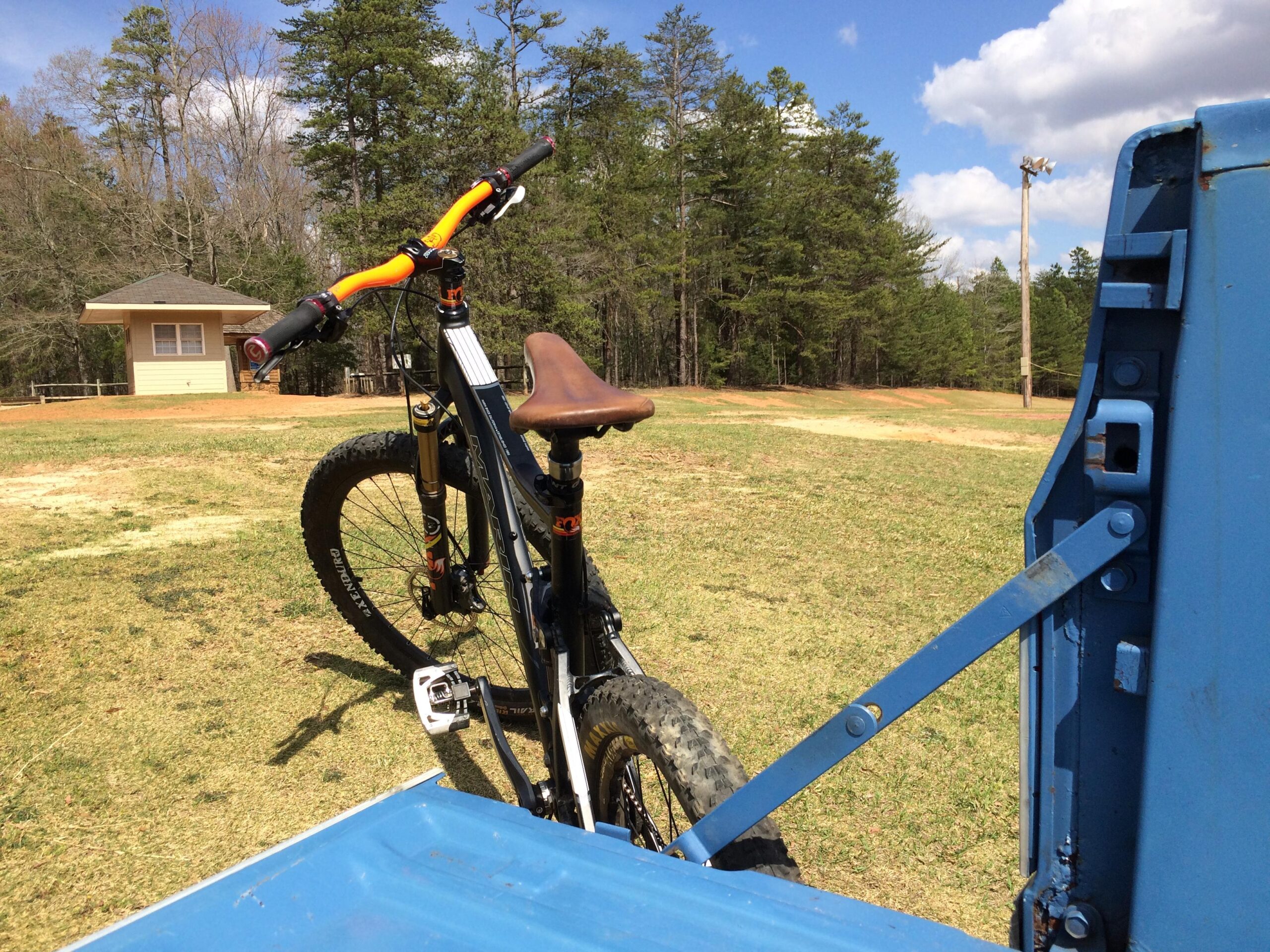 A mountain bicycle with orange handlebars parked next to a blue truck in a grassy area, framed by a background of trees and a small building under a partly cloudy sky. Anne Springs Close Greenway mountain bike trail.