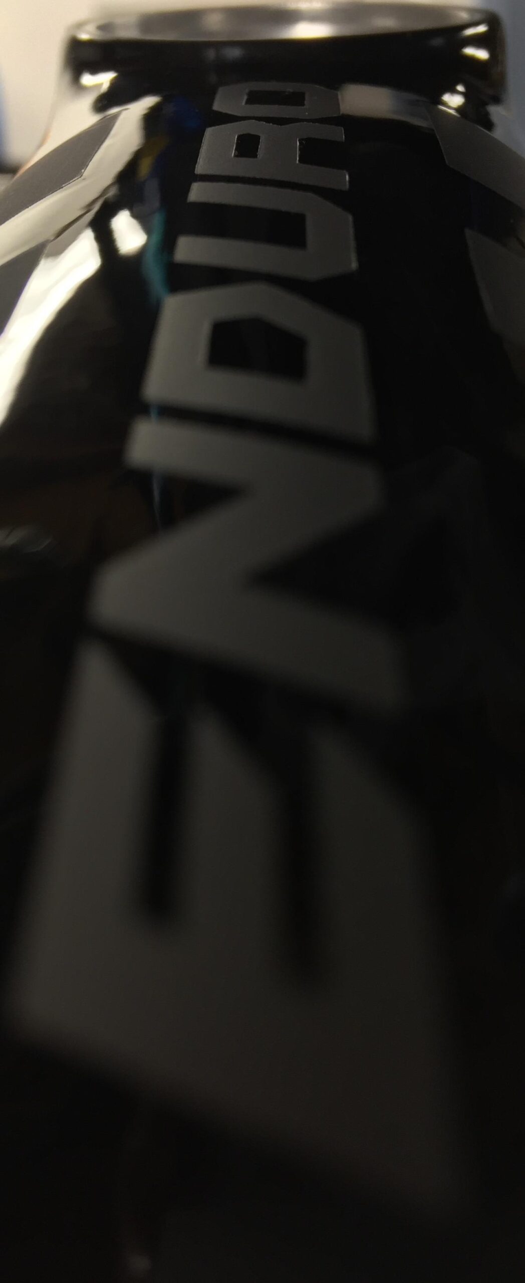 Specialized Enduro S-Works 650b: Close-up image of a shiny black object featuring bold, stylized lettering. The perspective emphasizes the glossy surface and captures light reflections, creating a dramatic effect. The background is blurred, adding focus to the object in the foreground.