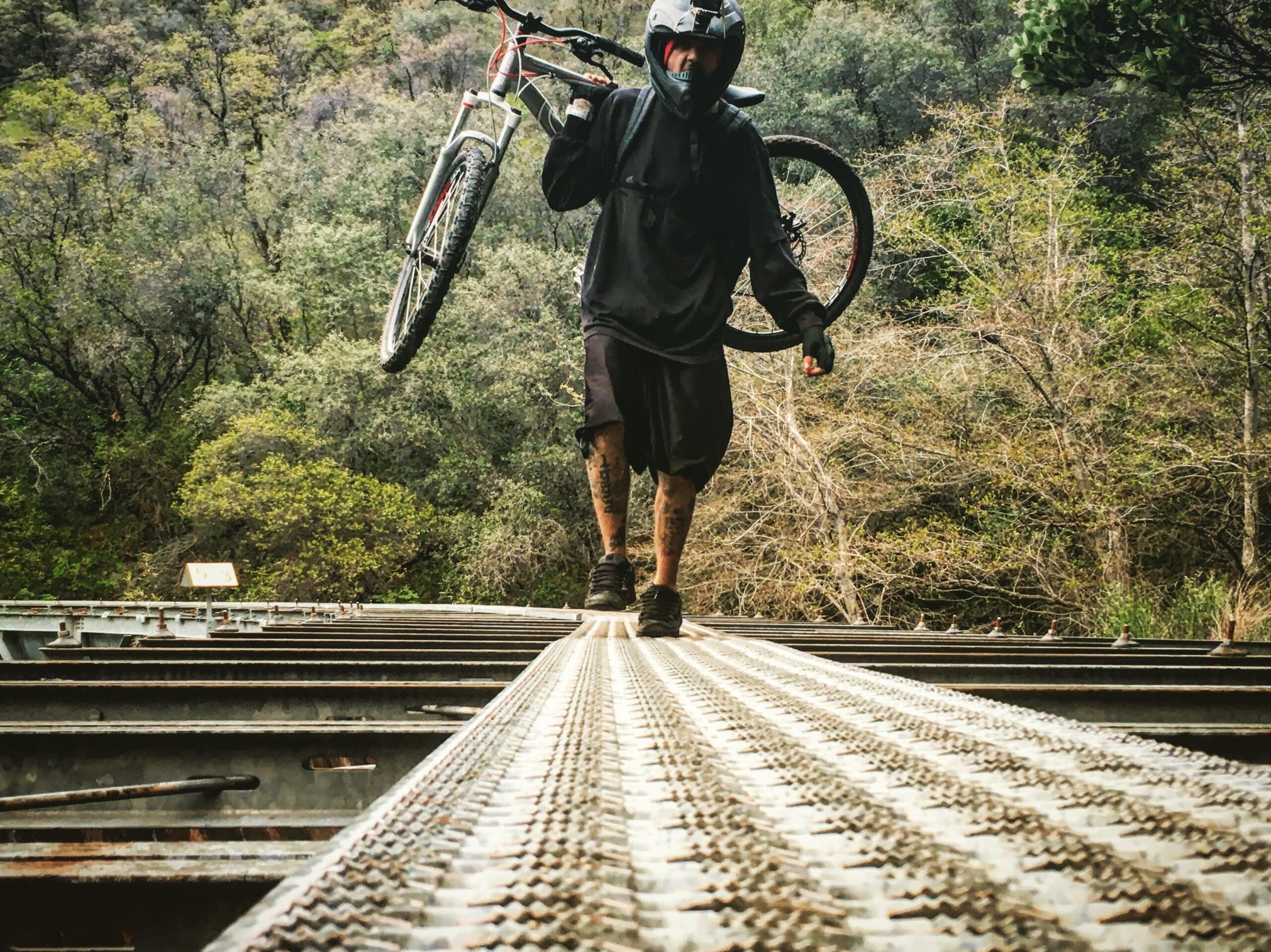 A cyclist dressed in dark clothing and a helmet walks on a metal bridge while carrying a mountain bike. The background features lush greenery, indicating a natural setting. The perspective emphasizes the height and structure of the bridge. Centerville Flumes mountain bike trail.
