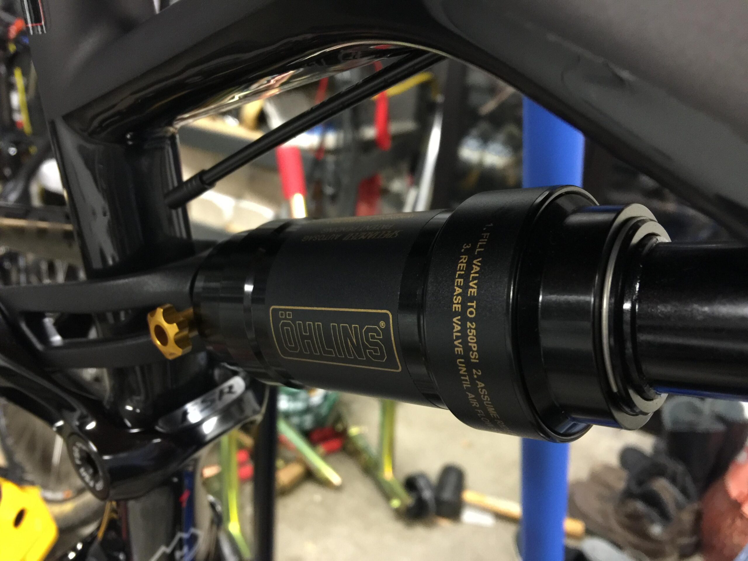 Specialized Enduro S-Works 650b: A close-up image of a black Öhlins bike shock absorber mounted on a bicycle frame, featuring a gold adjustment knob and visible technical specifications printed on the shock body. The background shows a mechanic's workspace with tools and bicycle parts.