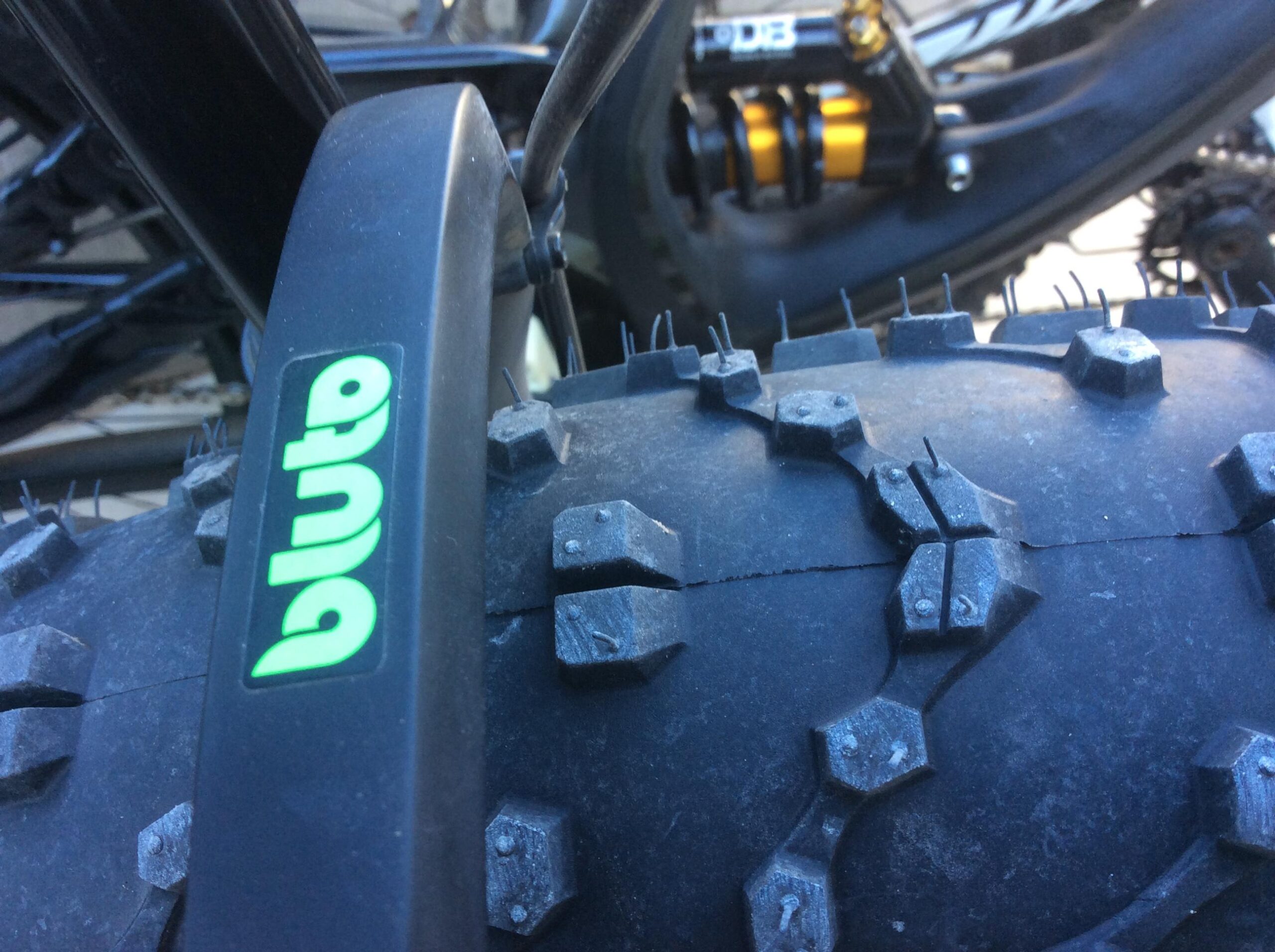 Close-up of a bicycle tire with a distinctive tread pattern and the logo "bluto" prominently displayed on the sidewall. The background features parts of a mountain bike frame and suspension components, suggesting an outdoor cycling environment.