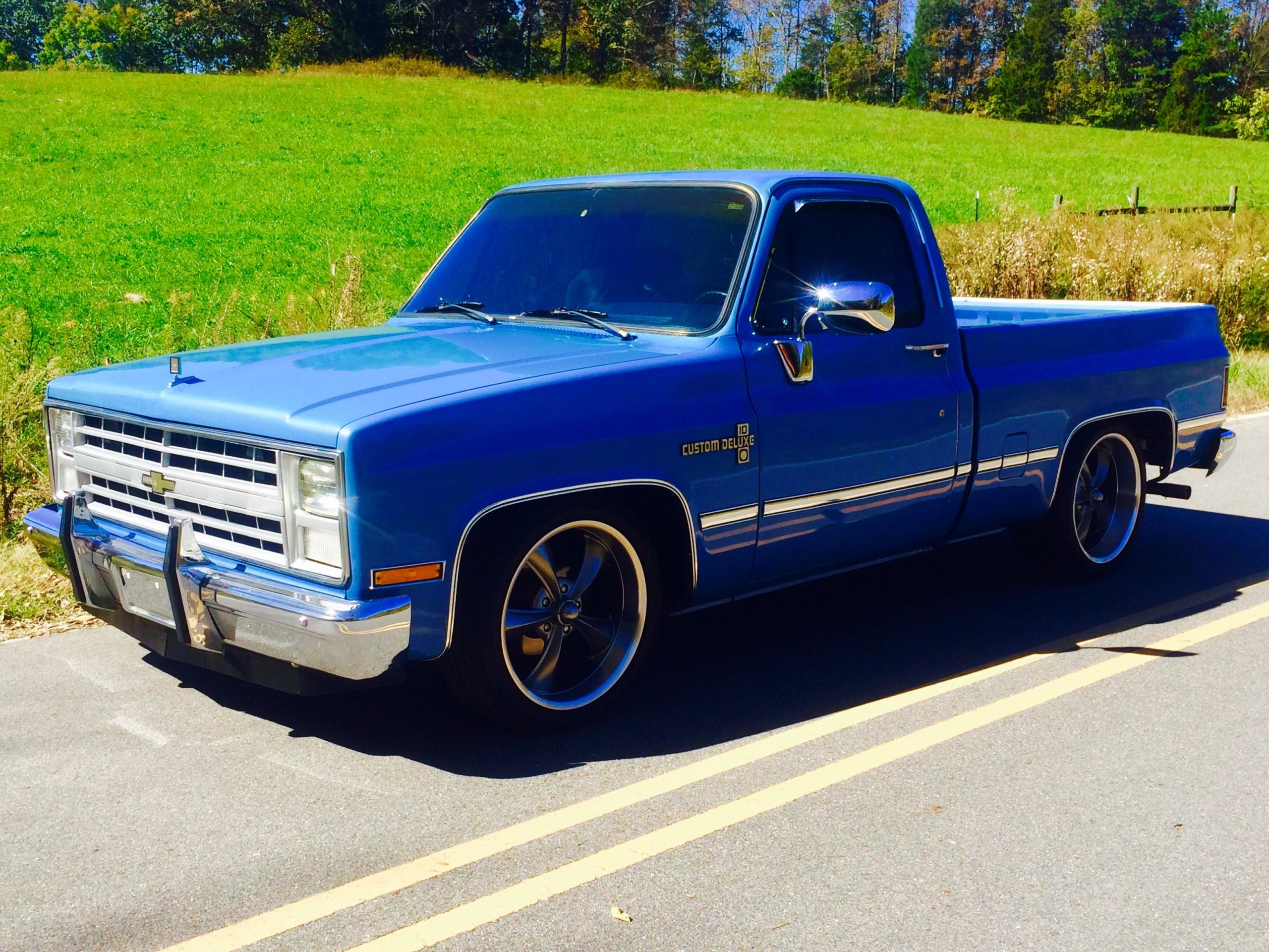 A vintage blue Chevrolet pickup truck is parked on the side of a road. It features a classic design, chrome accents, and sporty rims, with a lush green field and trees in the background. The truck is positioned at an angle, showcasing its front and side profile under bright sunlight. Warrior Creek mountain bike trail.