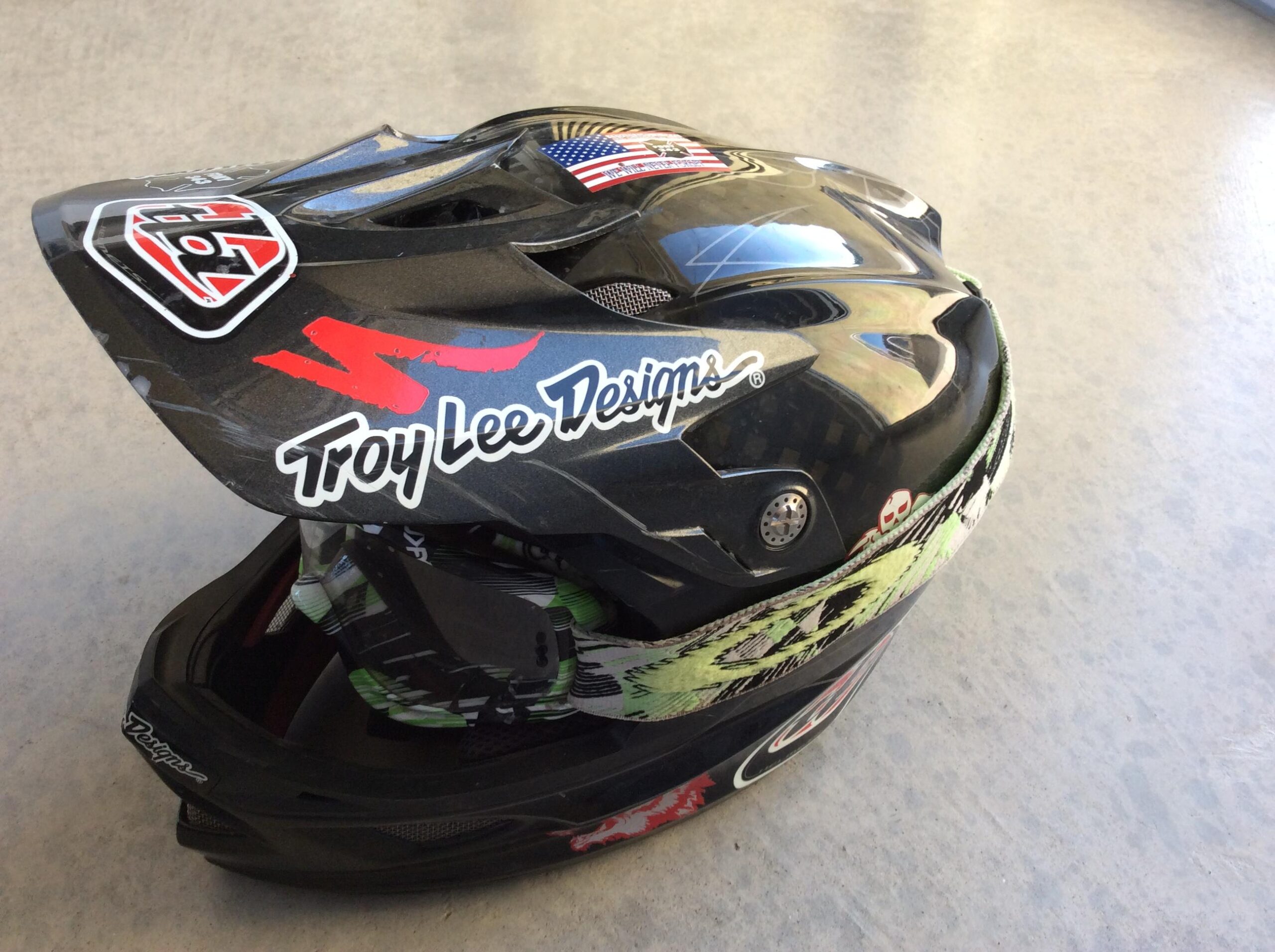 Black motocross helmet featuring Troy Lee Designs logo, American flag sticker, and a green patterned strap. The helmet has various decorative graphics and patterns and is positioned on a light concrete surface.