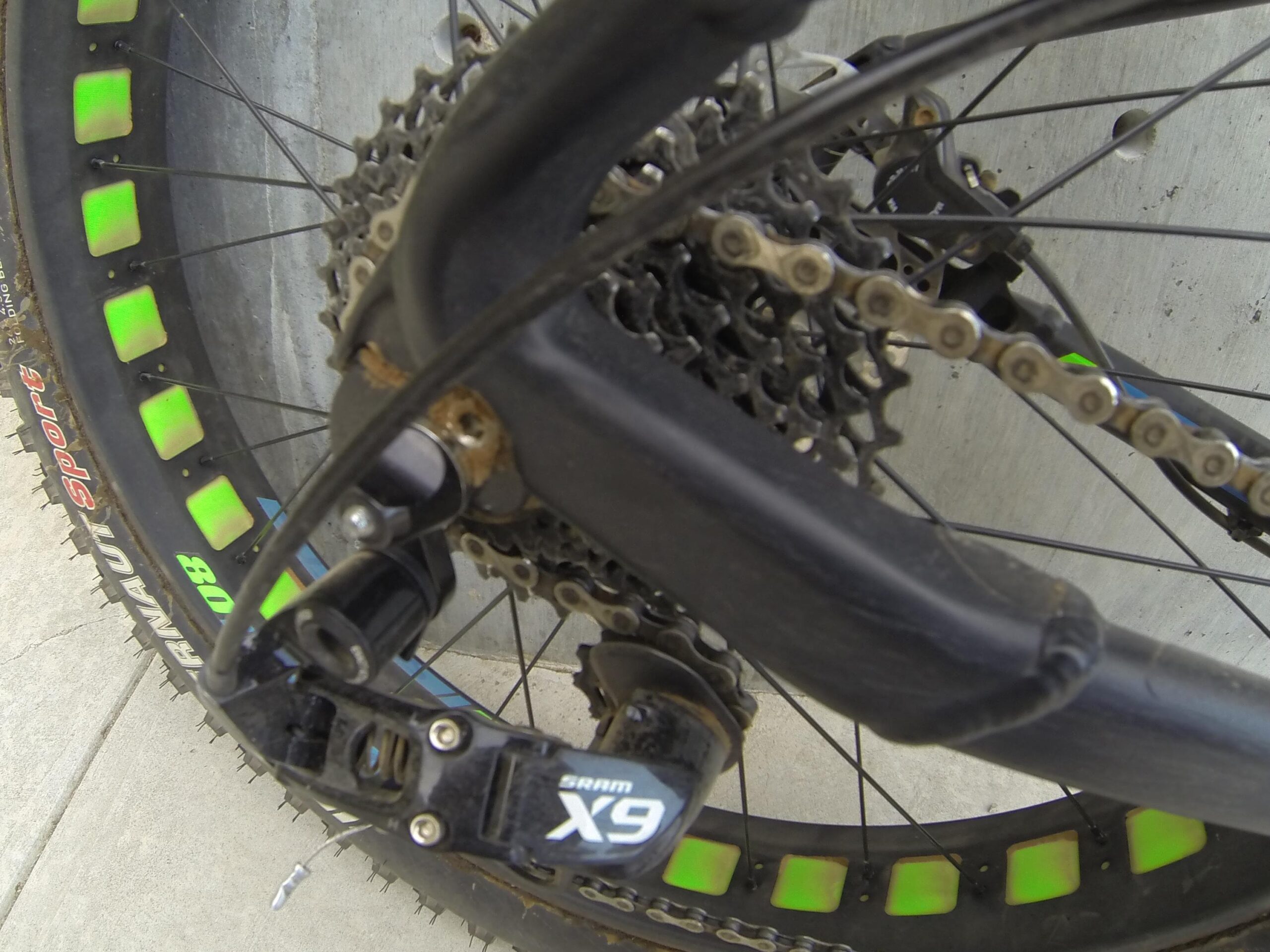 Scott Big Ed: Close-up image of a bicycle's rear wheel and drivetrain. The photo shows the cassette, chain, and derailleur, with a focus on the SRAM X9 components. The tire features bright green reflective accents. The background consists of a concrete surface.
