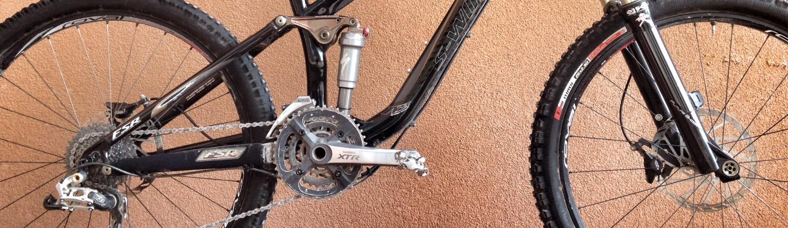 Specialized S-Works Enduro SL Carbon: A close-up view of a black mountain bike featuring a rear suspension system, a crankset with Shimano XTR components, and detailed chain and gear mechanisms. The bike is positioned against a textured, neutral-colored wall.