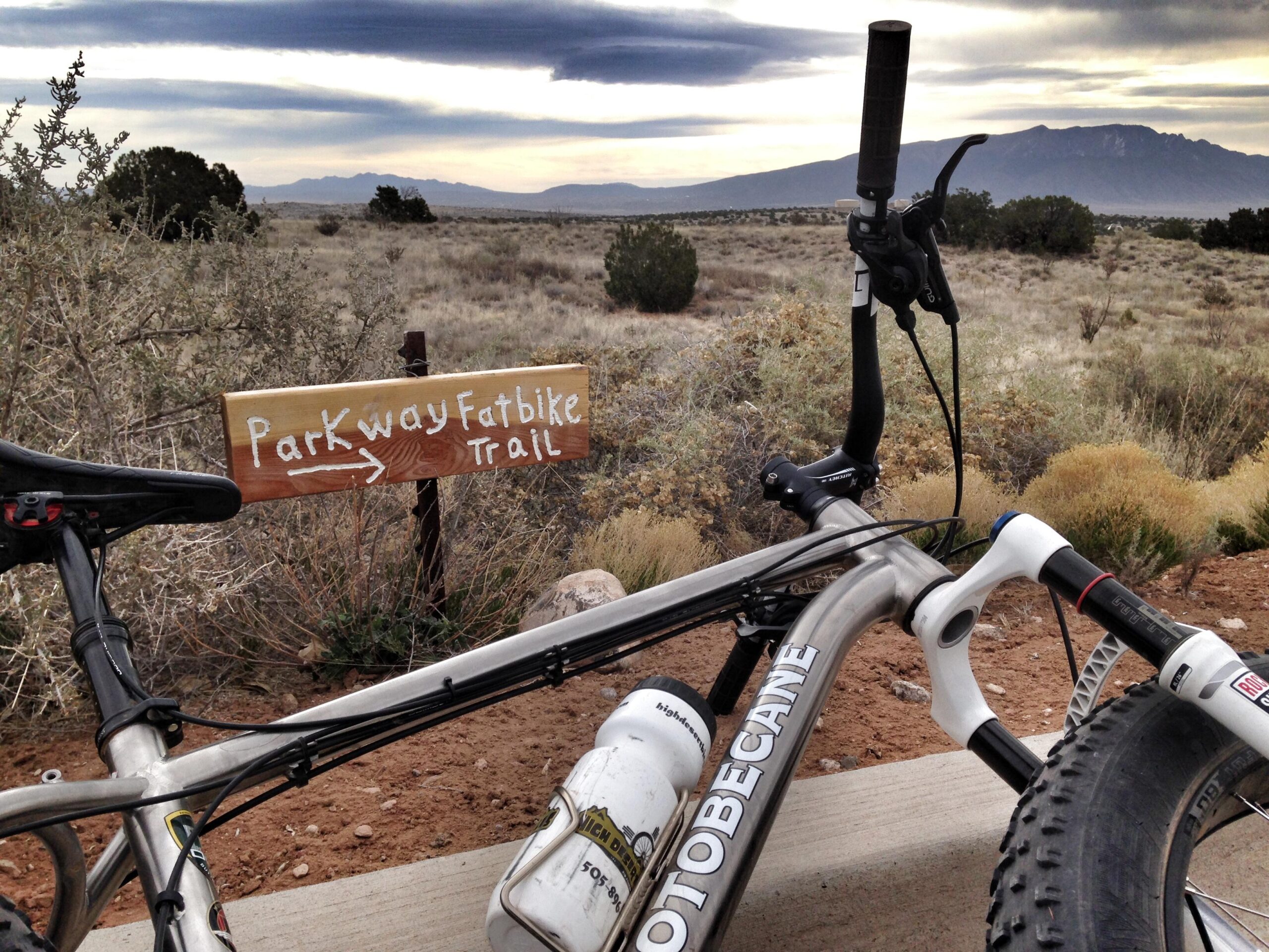 A close-up view of a fat tire bike parked near a wooden trail sign that reads "Parkway Fatbike Trail." The bike is positioned on a dirt path with a landscape of shrubs and distant mountains under a cloudy sky. Parkway Fatbike trail mountain bike trail.