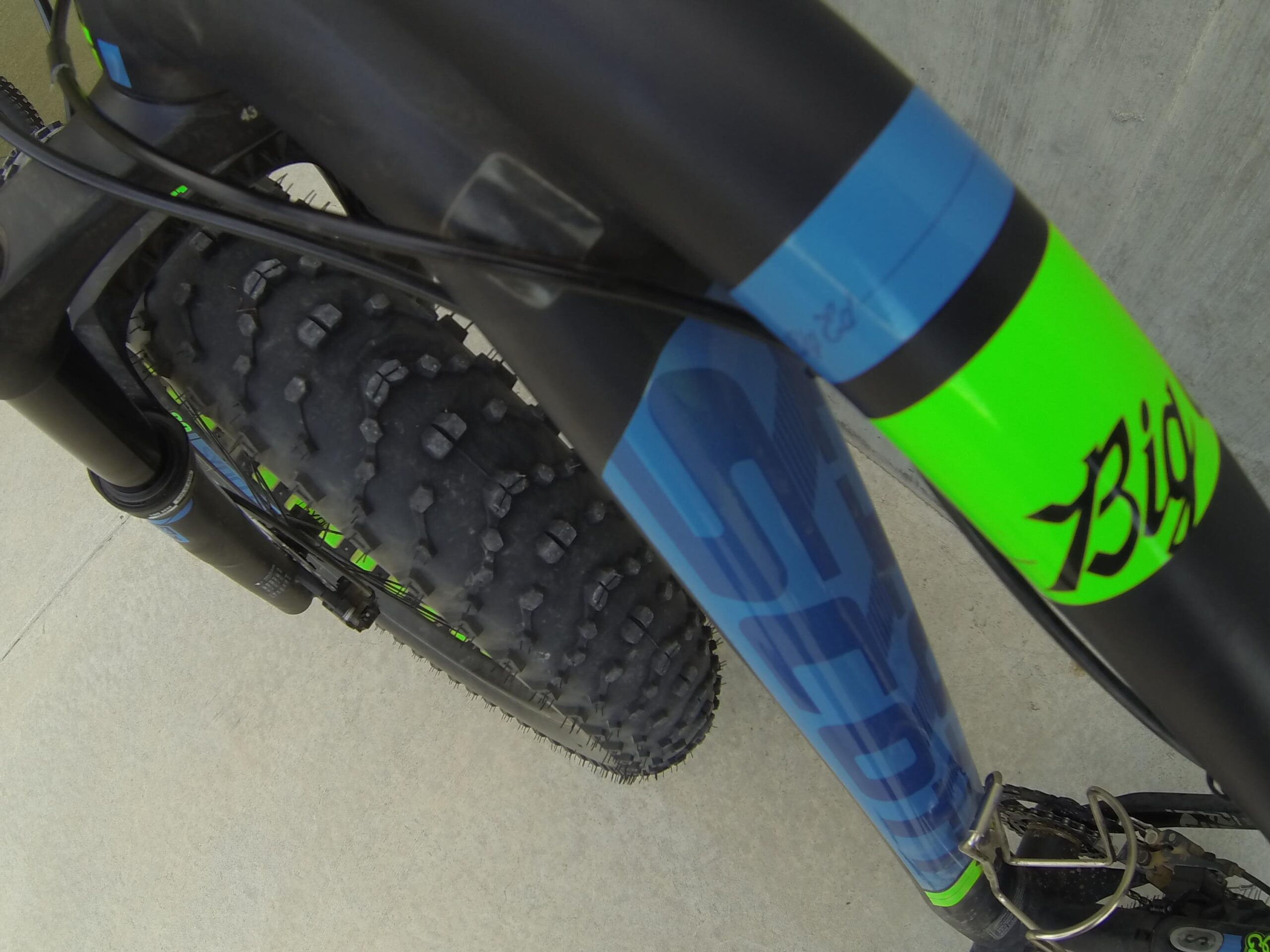 Scott Big Ed: Close-up view of the front fork and tire of a mountain bike, featuring a mix of black, blue, and bright green colors. The tire displays a rugged tread pattern, suitable for off-road terrain. The image captures the bike's branding and design details.