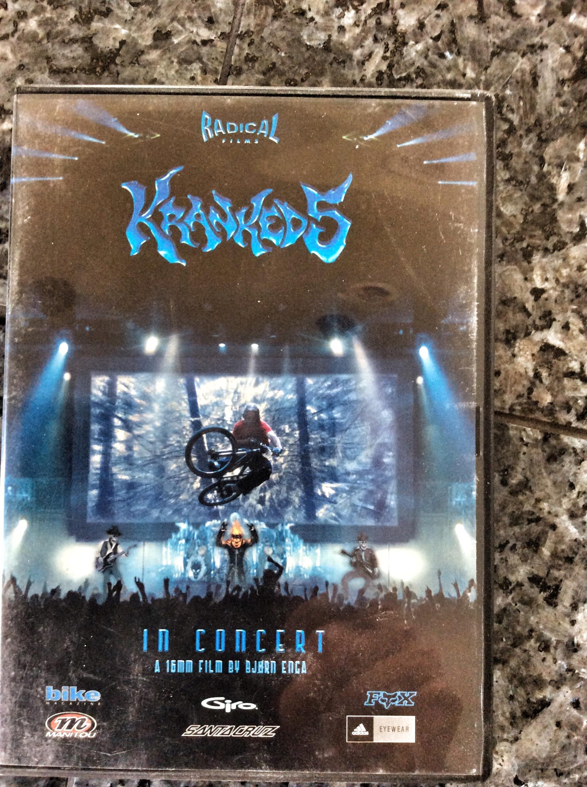 DVD cover for "Kranked 5," showing a BMX rider performing a stunt in front of an audience and a large screen displaying a forest scene. The title "Kranked 5" is prominently displayed in blue lettering, with the tagline "In Concert" below, along with credits for the film and various sponsor logos. The background features a concert atmosphere with lights and silhouettes of musicians.