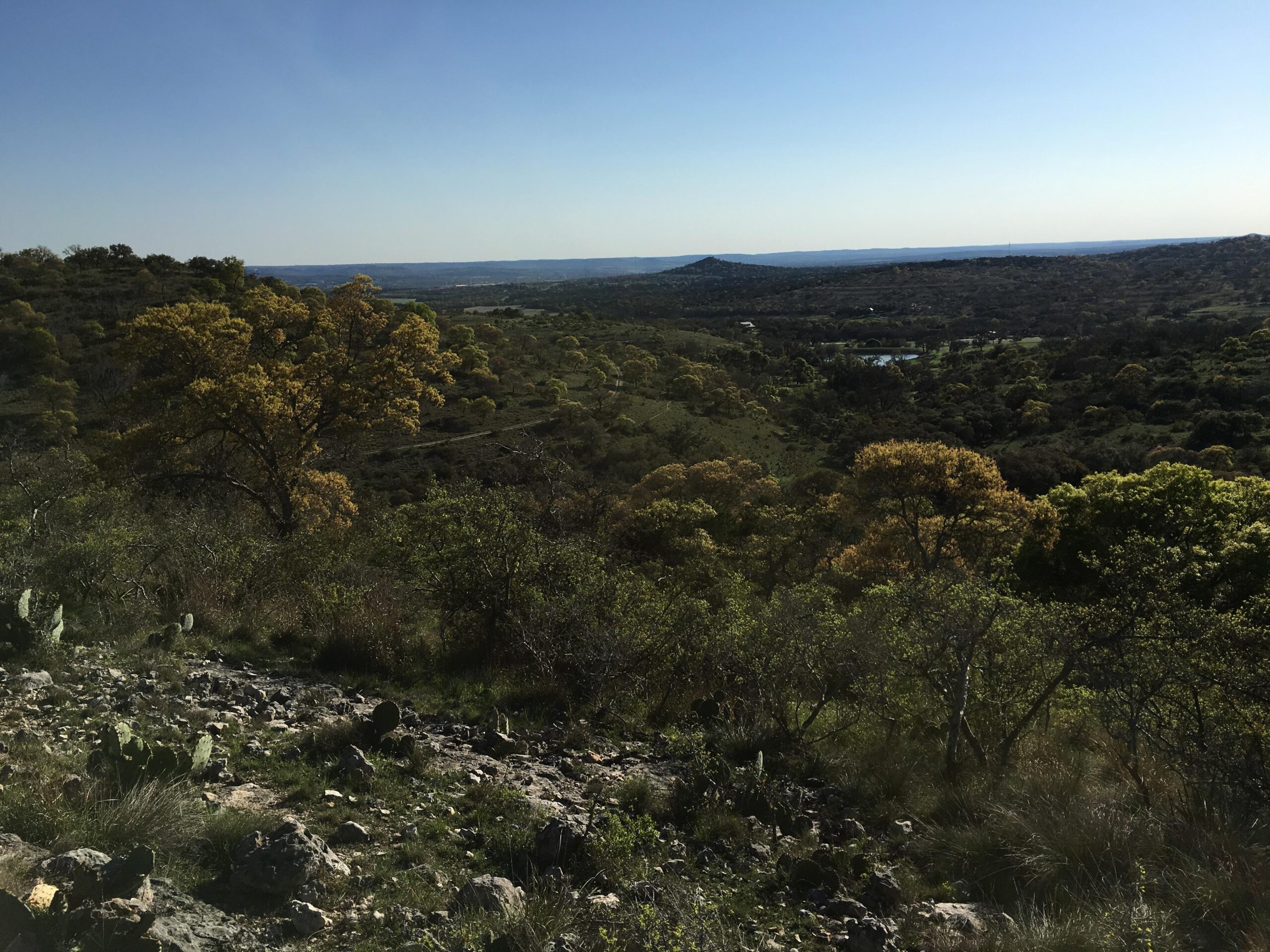 A panoramic view of rolling hills covered in greenery, with trees displaying hints of yellow foliage. The landscape features rocky terrain in the foreground and a distant horizon under a clear blue sky. A small body of water can be seen nestled among the trees in the middle ground. Flat Rock Ranch mountain bike trail.