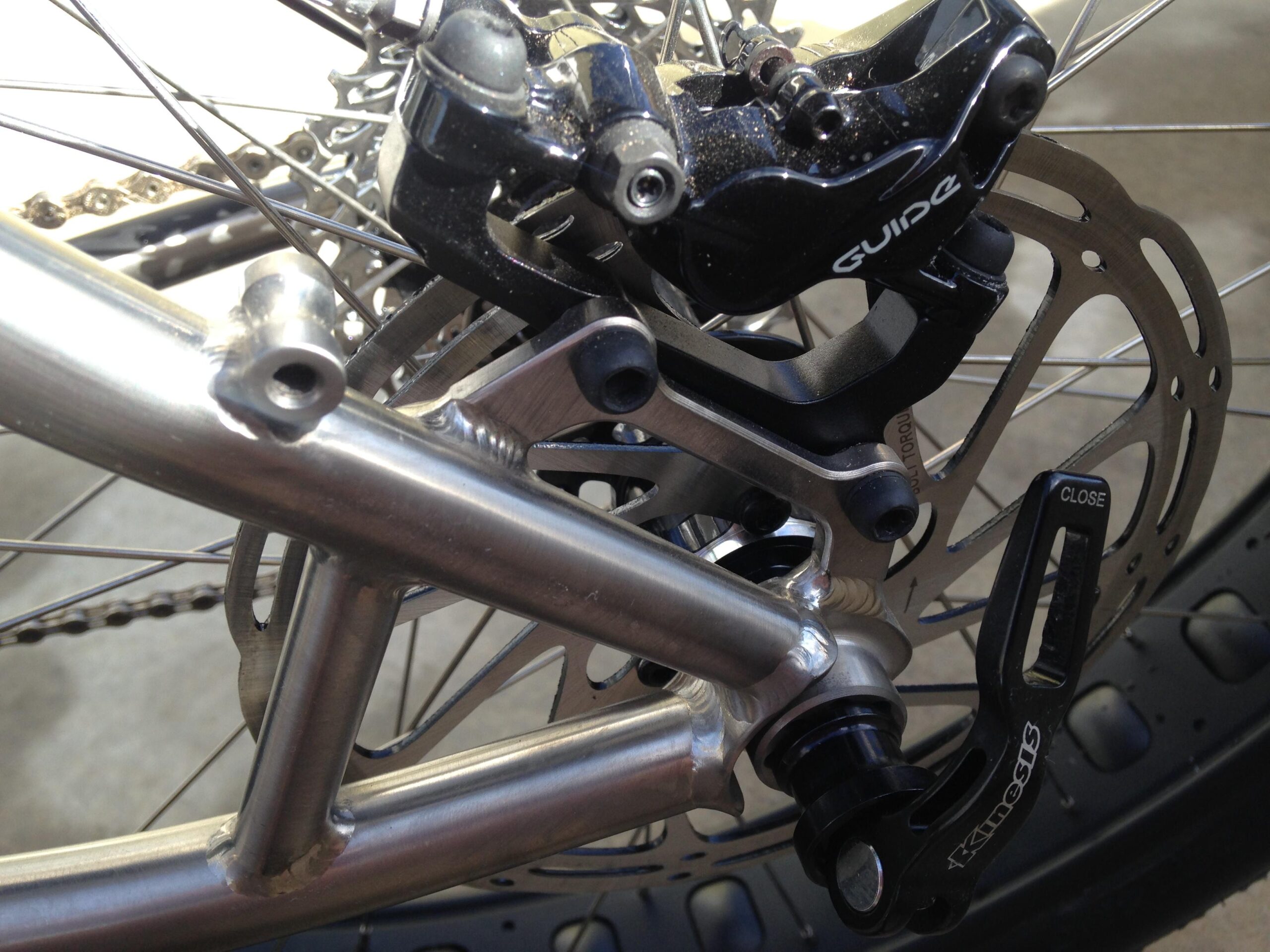 Motobecane Motobecane Bullet Titanium Fat bike: Close-up view of a bicycle's rear brake system, featuring a metallic frame, disc brake, and a black brake caliper labeled "Guide." The image highlights the intricate components, including the brake rotor and part of the bicycle's drivetrain.