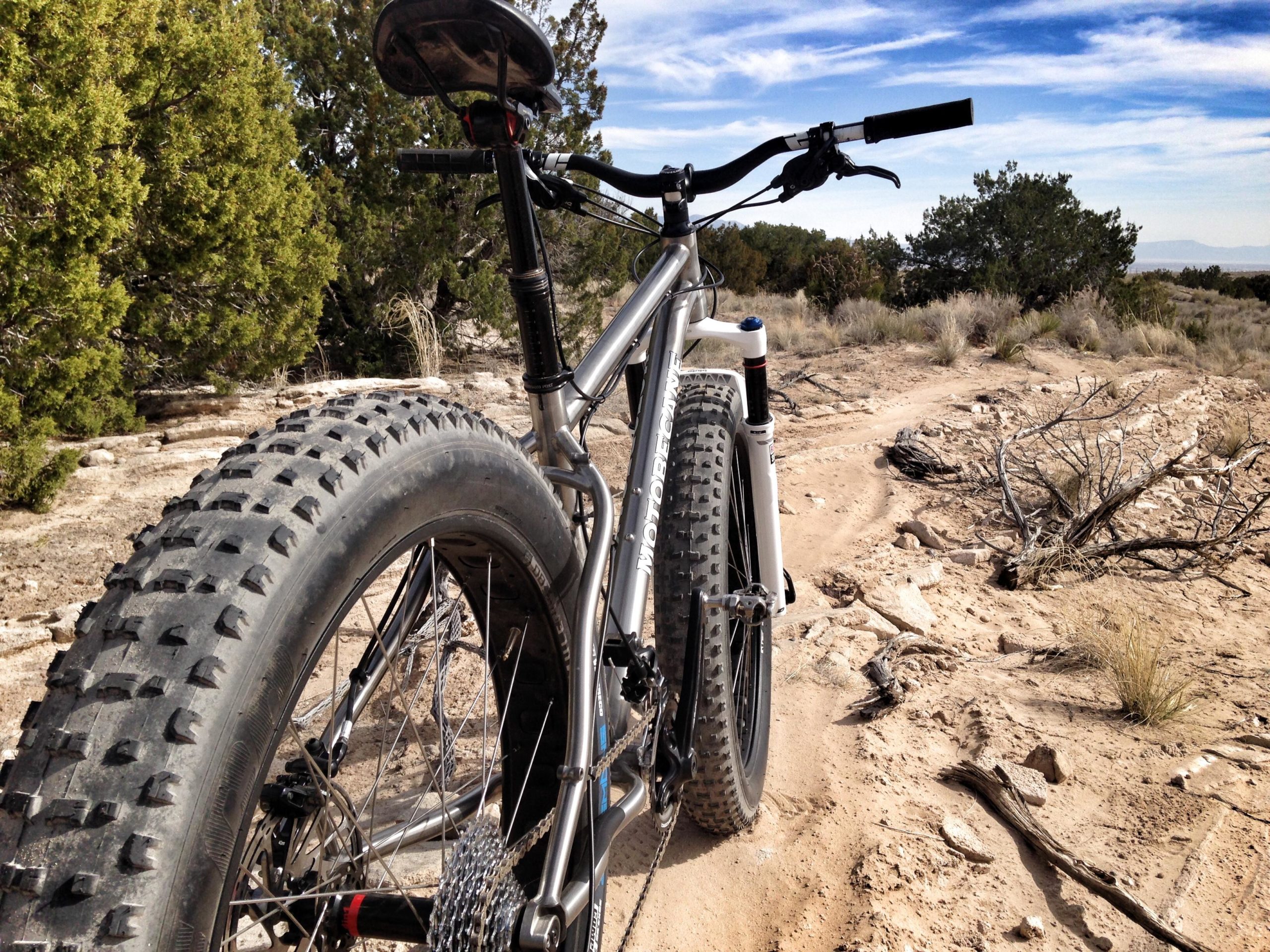 A close-up view of a mountain bike with large, knobby tires, parked on a sandy trail surrounded by bushes and sparse trees under a blue sky with clouds. Parkway Fatbike trail mountain bike trail.
