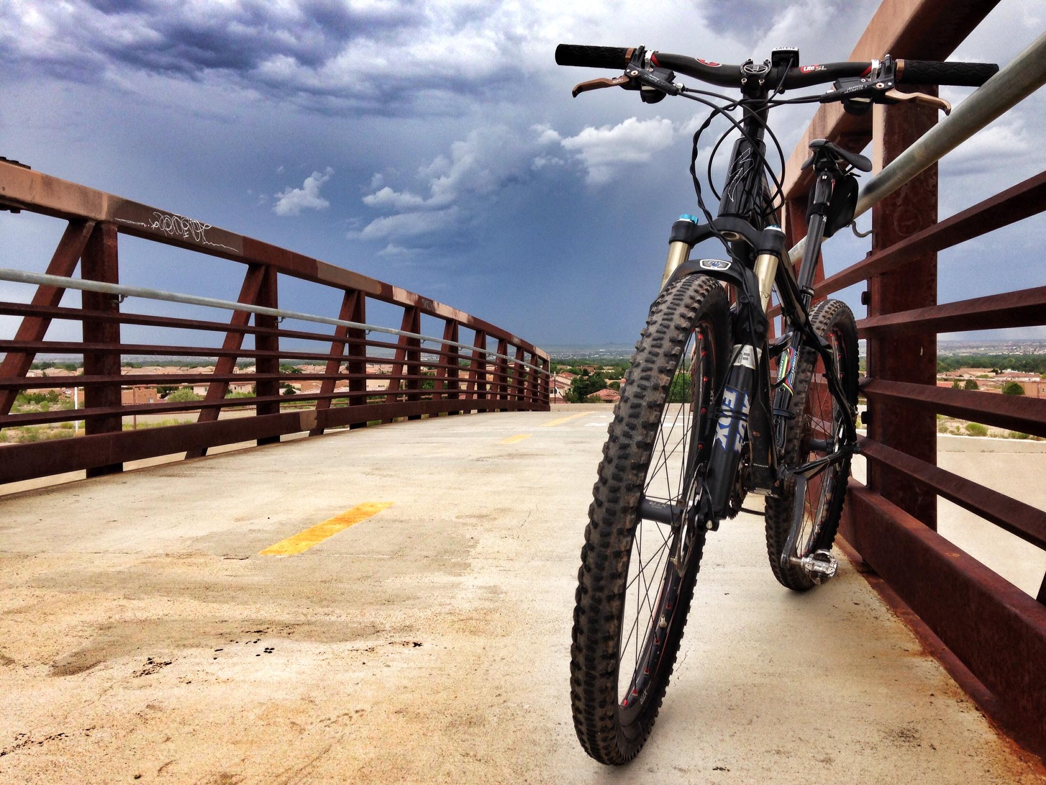 A mountain bike resting on a bridge, with a metal railing and a cloudy sky in the background. The bridge has a smooth, light-colored surface with a faint yellow line visible. The landscape beyond shows a mix of greenery and buildings, suggesting an urban setting. The overall mood is calm, with the impending clouds hinting at possible rain. Albuquerque Bosque mountain bike trail.