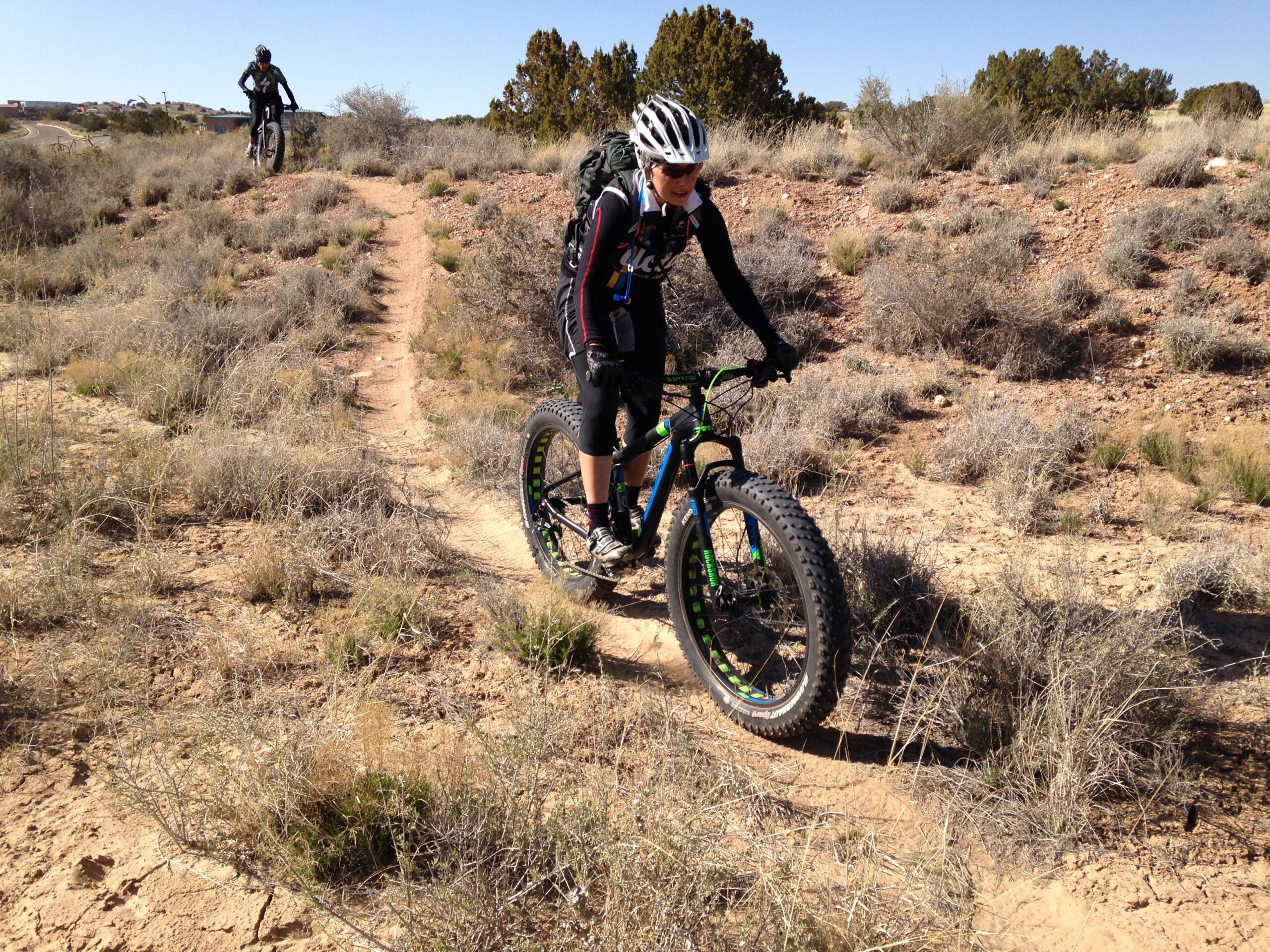 A cyclist riding a fat-tire bike along a dirt trail in a desert landscape, surrounded by sparse vegetation and shrubs. In the background, another cyclist is navigating a similar trail, with a clear blue sky overhead. Super Fat Bike Loop mountain bike trail.