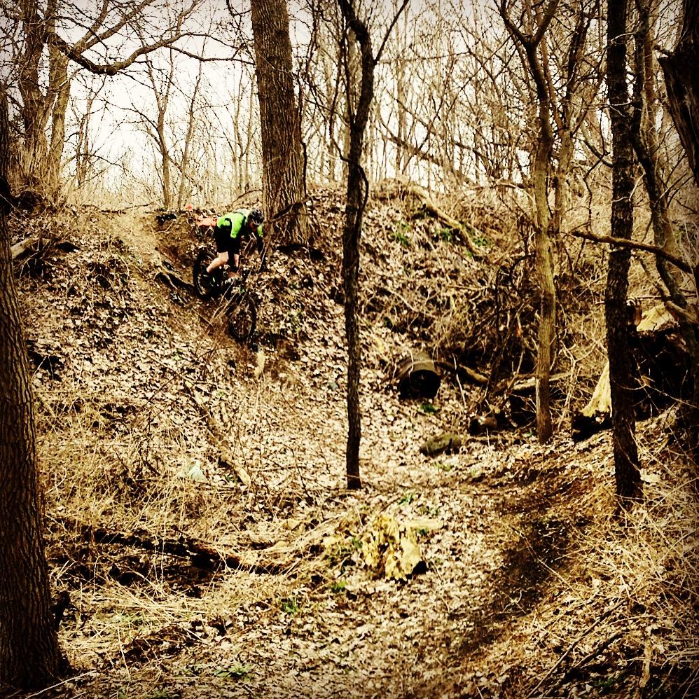 A cyclist in a bright green shirt navigates a rocky trail in a wooded area, surrounded by bare trees and fallen leaves. The scene depicts a challenging terrain with a hillside and a winding path, capturing the spirit of outdoor adventure. The Dump mountain bike trail.