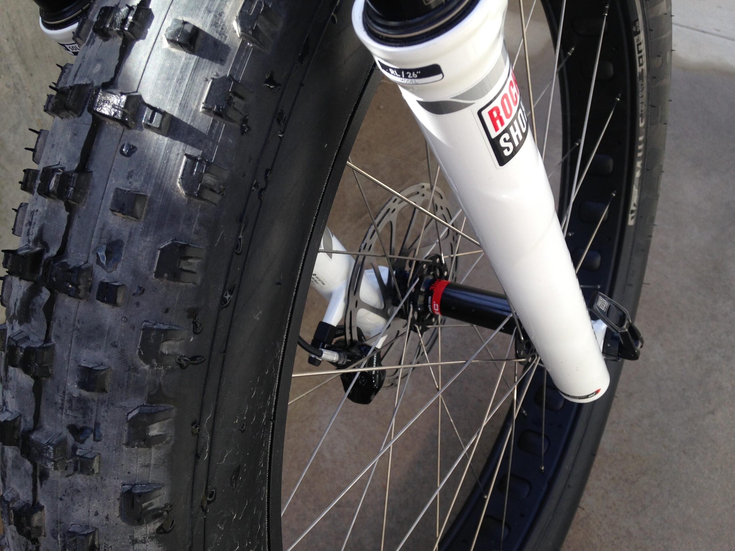 Motobecane Motobecane Bullet Titanium Fat bike: Close-up view of a bicycle's front wheel featuring a thick, knobby tire, designed for off-road use. The bike's suspension fork is visible in white, highlighting the details of the wheel hub and disc brake. The background shows a concrete surface.
