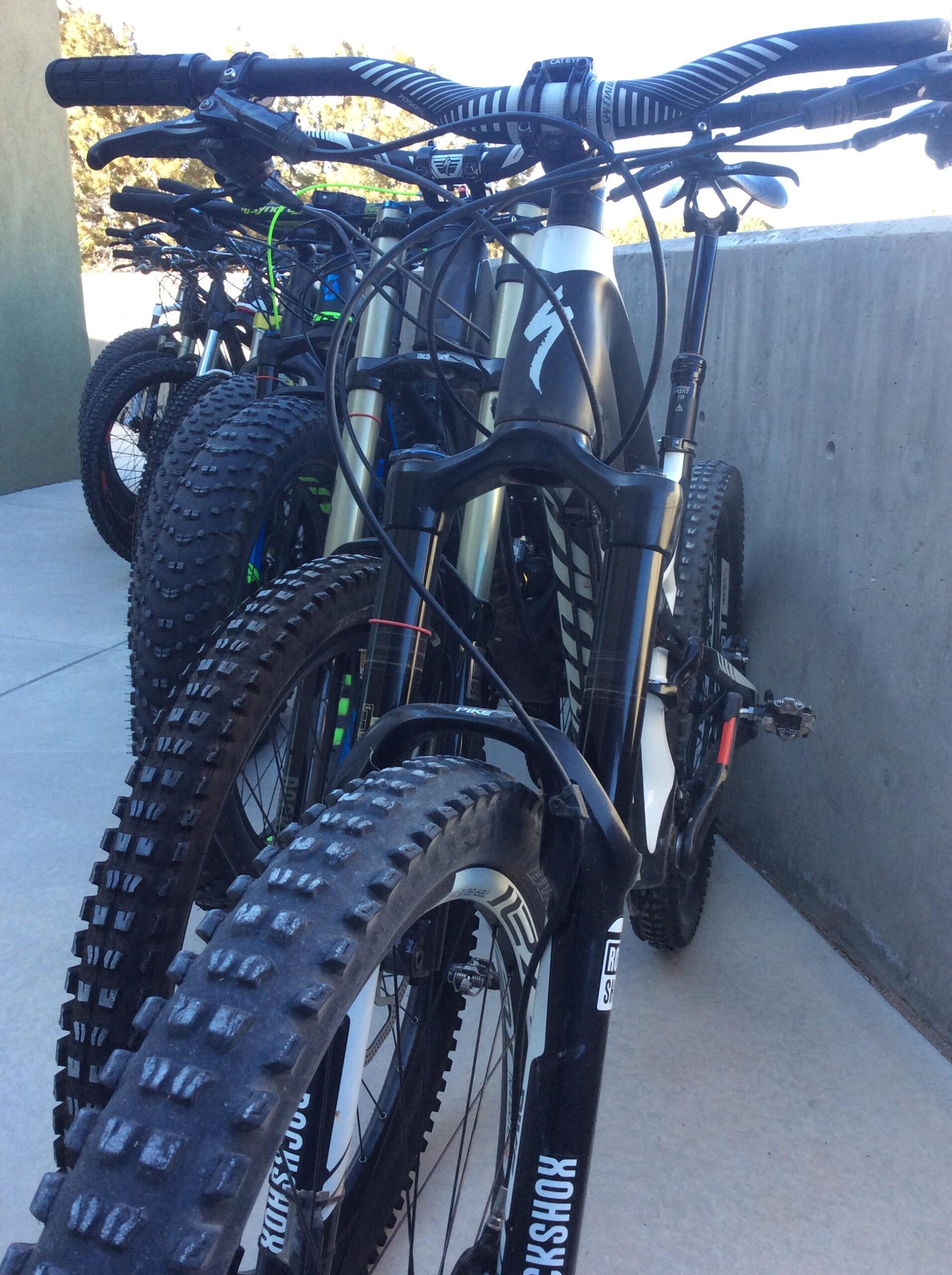 A row of mountain bikes parked against a concrete wall. The image focuses on the front ends of the bikes, showcasing their thick, knobby tires and handlebars. The bikes feature various colors and designs, indicating they're designed for off-road riding. The setting has a bright, outdoor atmosphere.