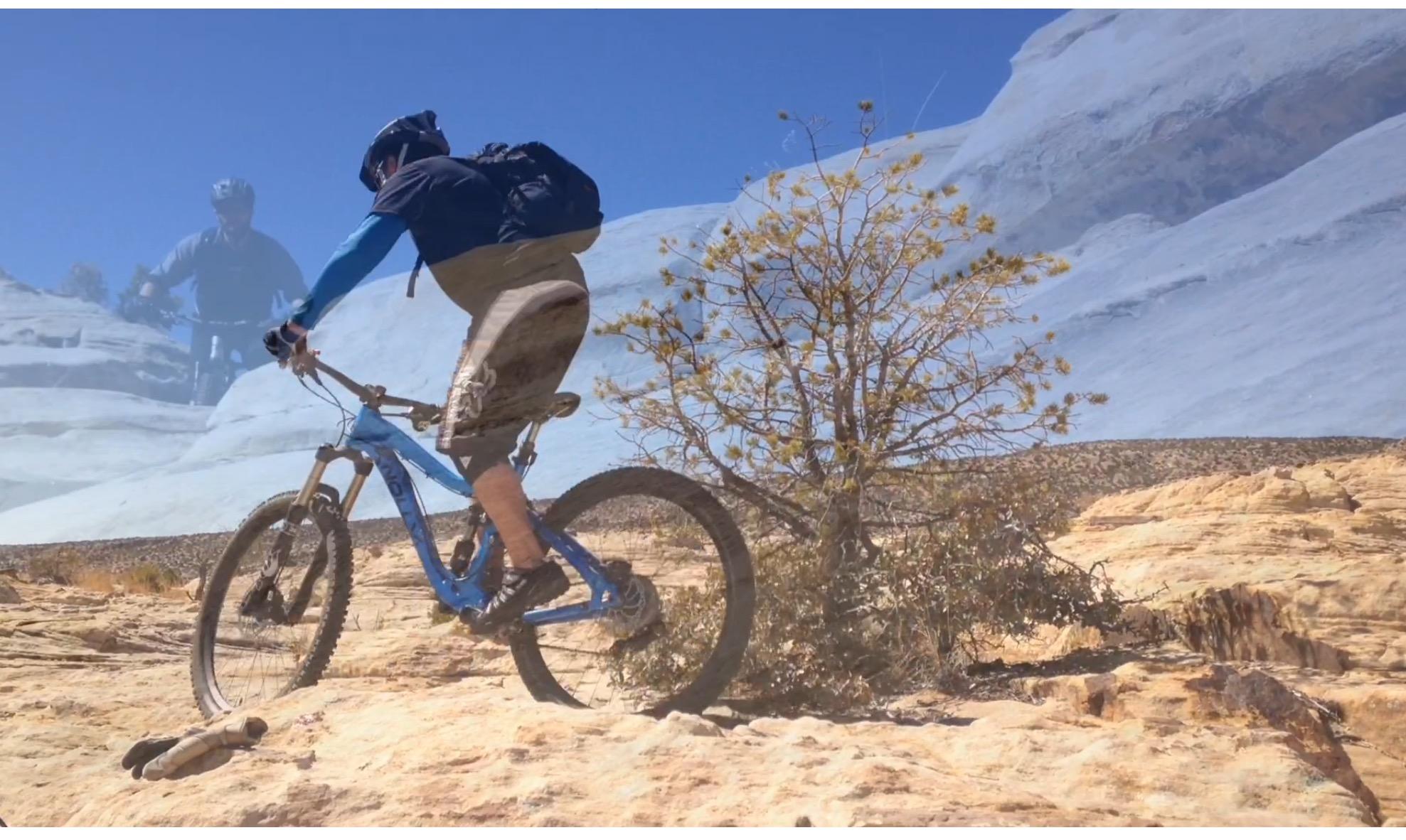 Knolly Chilcotin: A mountain biker riding over rocky terrain with a blue sky in the background. A small tree is visible nearby, and there are some scattered shrubs and natural formations in the landscape. The biker is wearing a helmet and a blue shirt, showcasing an action-oriented outdoor activity.