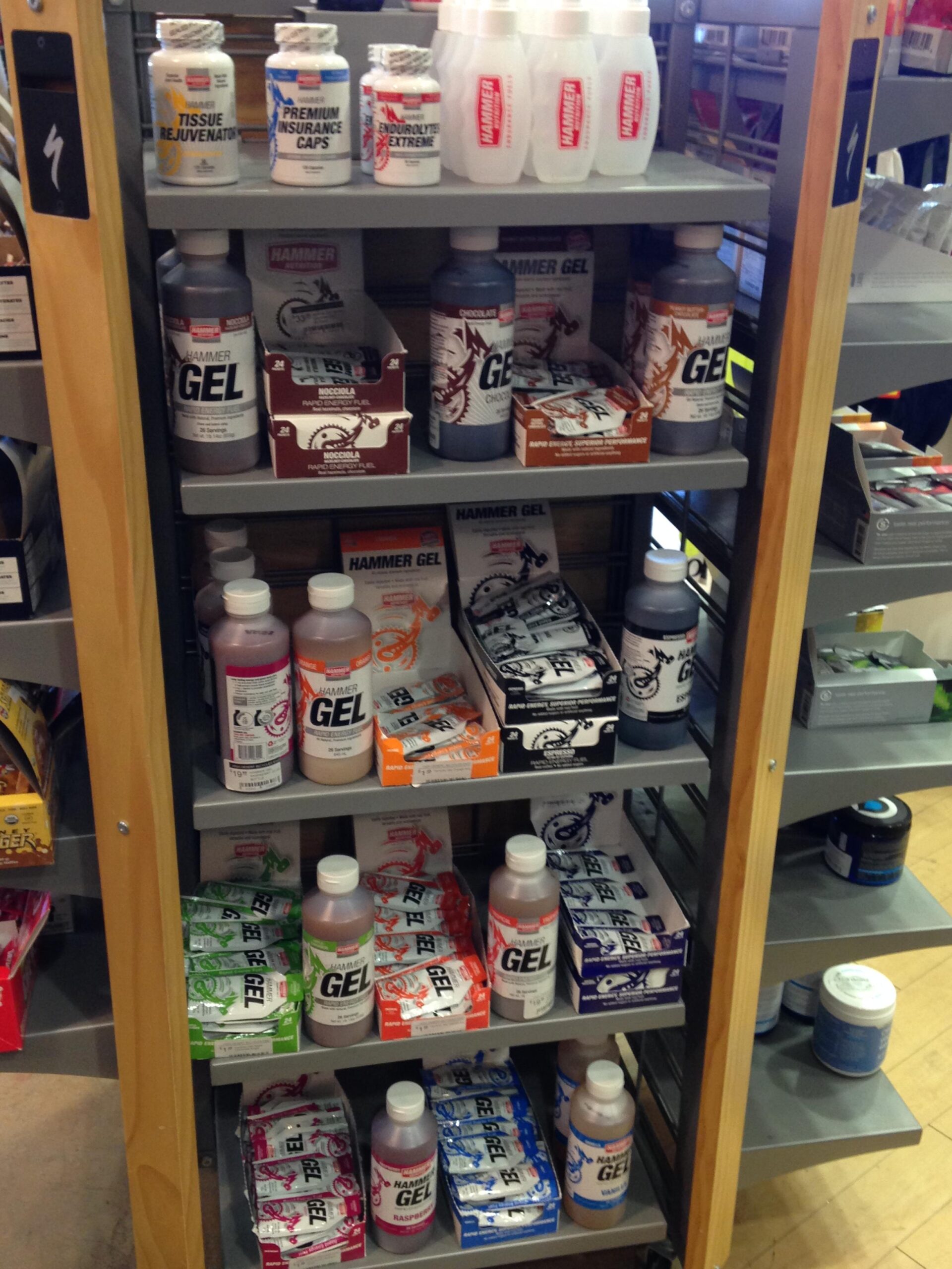 A display shelf in a store featuring various sports nutrition products, including Hammer Gel bottles and packets in multiple flavors, along with other supplements like tissue rejuvenation caps and endurance products.