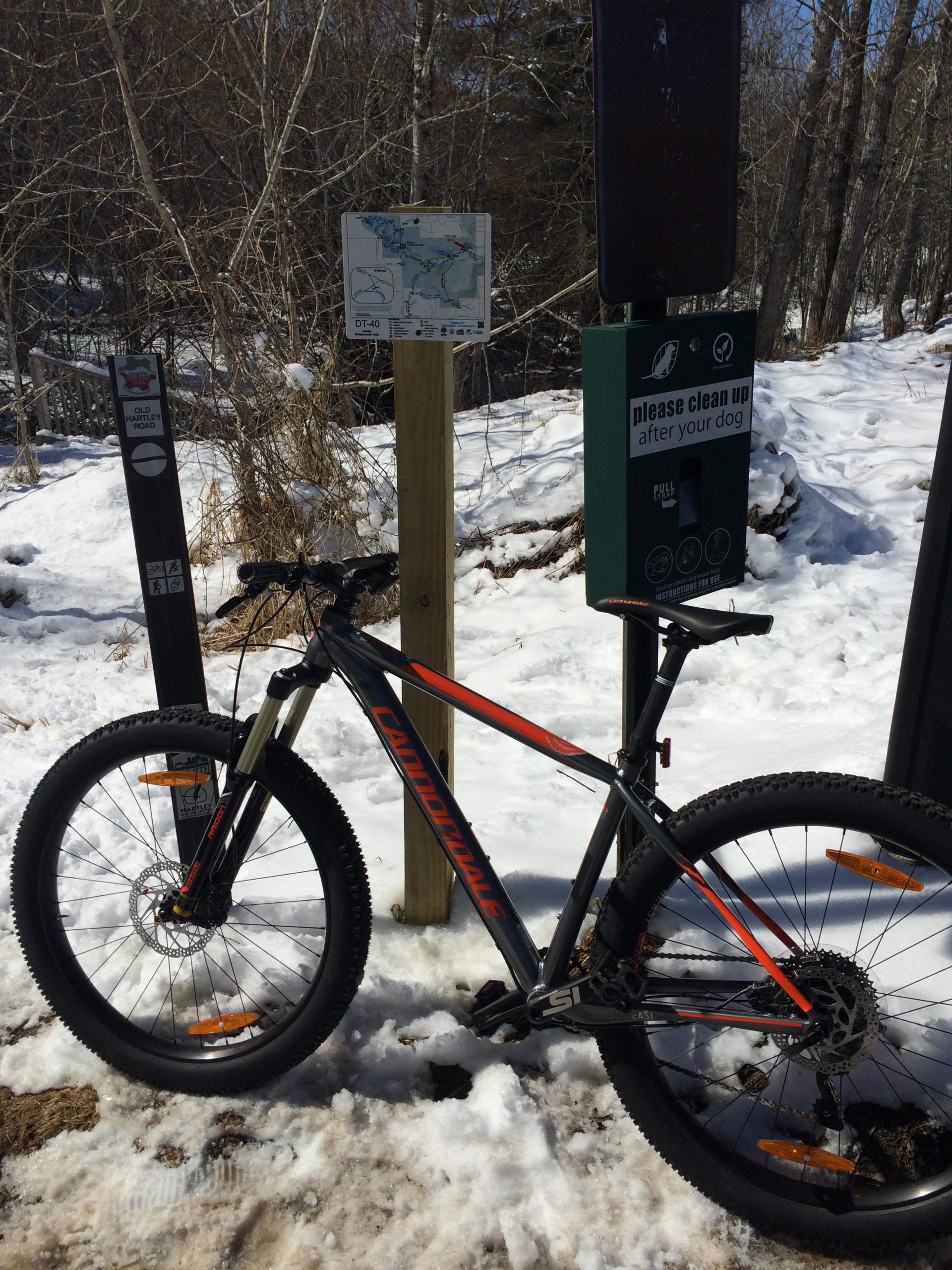 A mountain bike resting on snowy ground near a trail sign with a map and dog waste station. Trees are visible in the background. Hartley Park mountain bike trail.